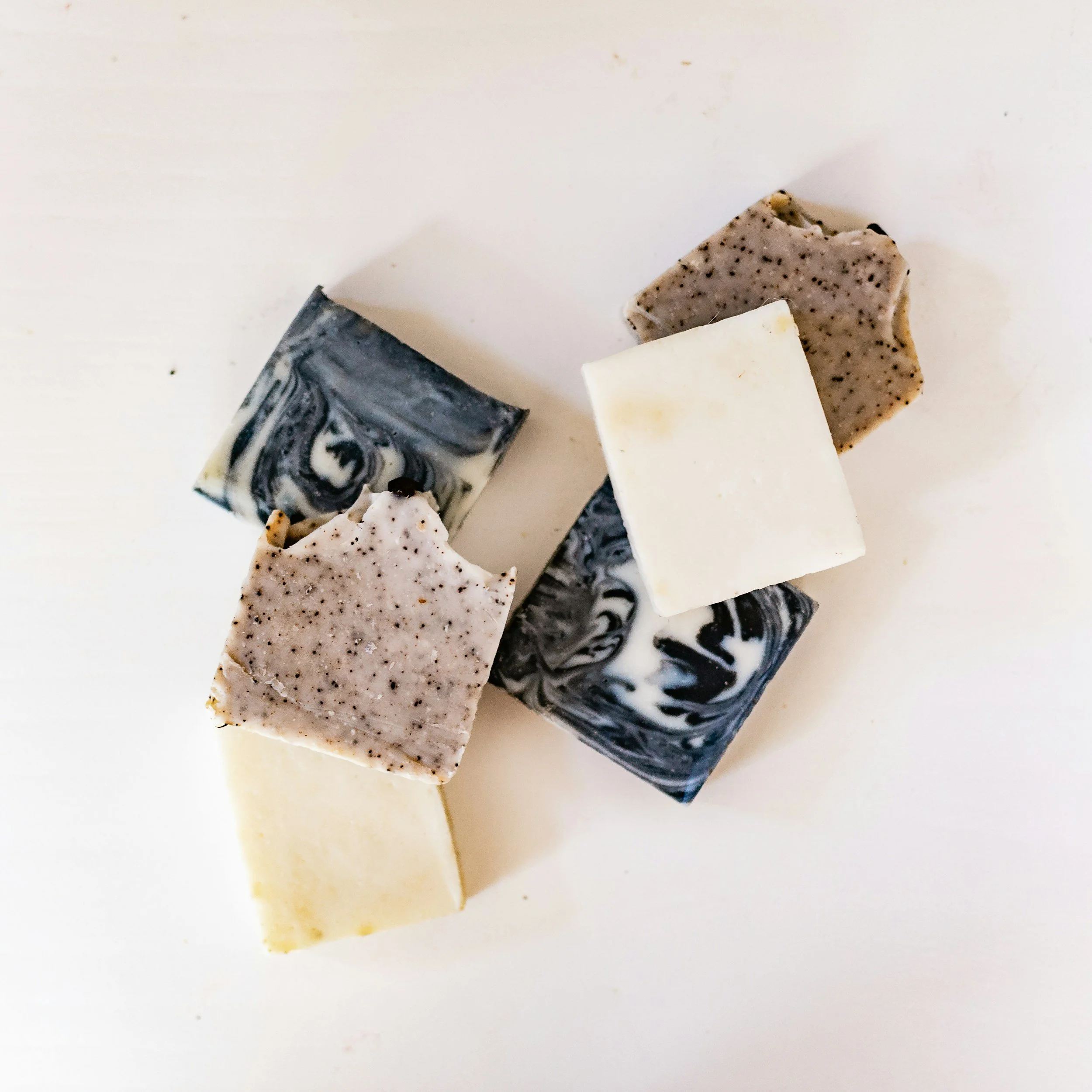 A collection of various types of soap bars, including black and white marbled soap, speckled beige soap, and plain white soap, arranged on a white surface.