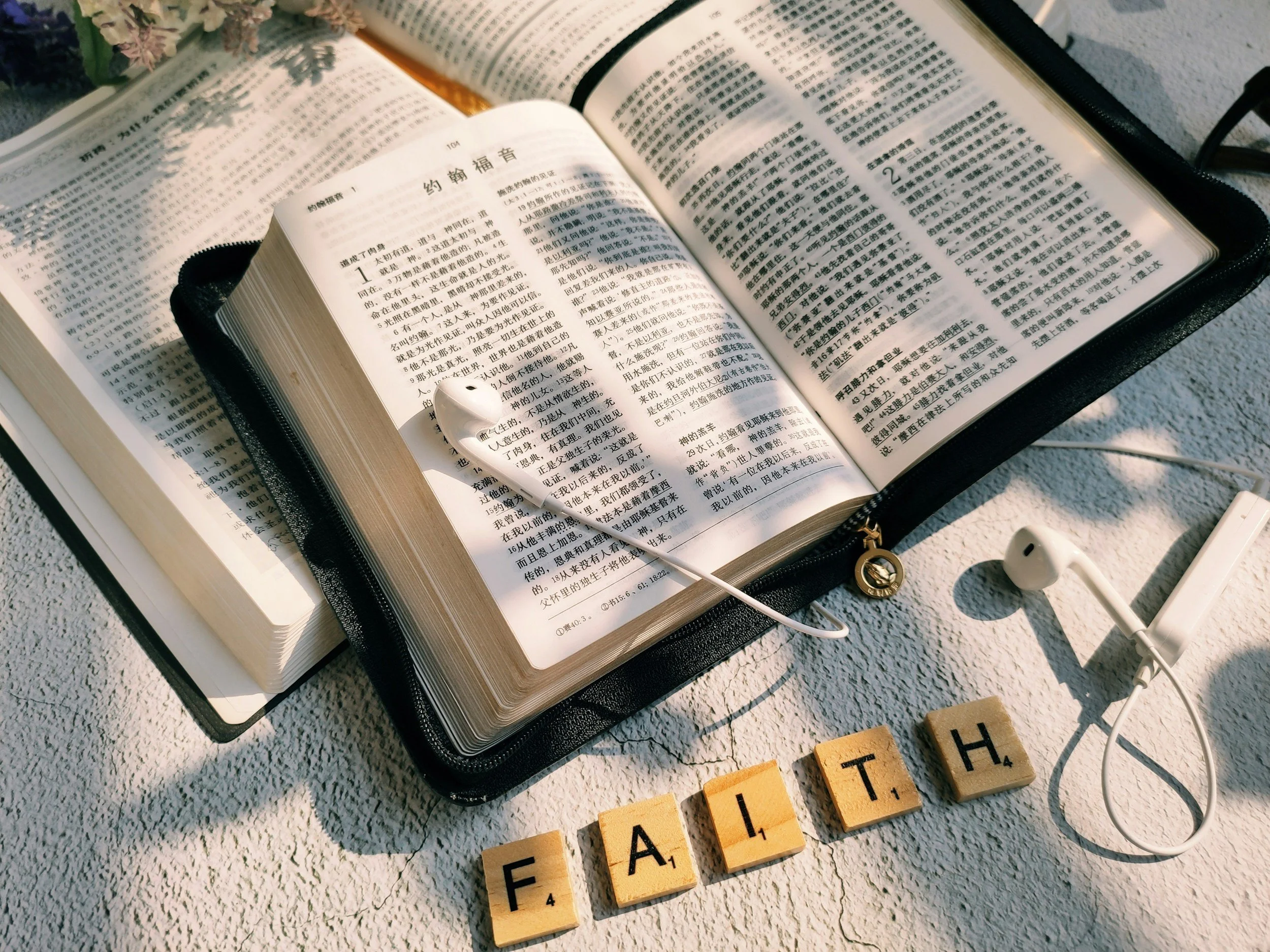 Open Bible in Chinese with olive wood letter tiles spelling 'FAITH', white earphones, small bricks, and a white device on a textured surface with sunlight and shadows.