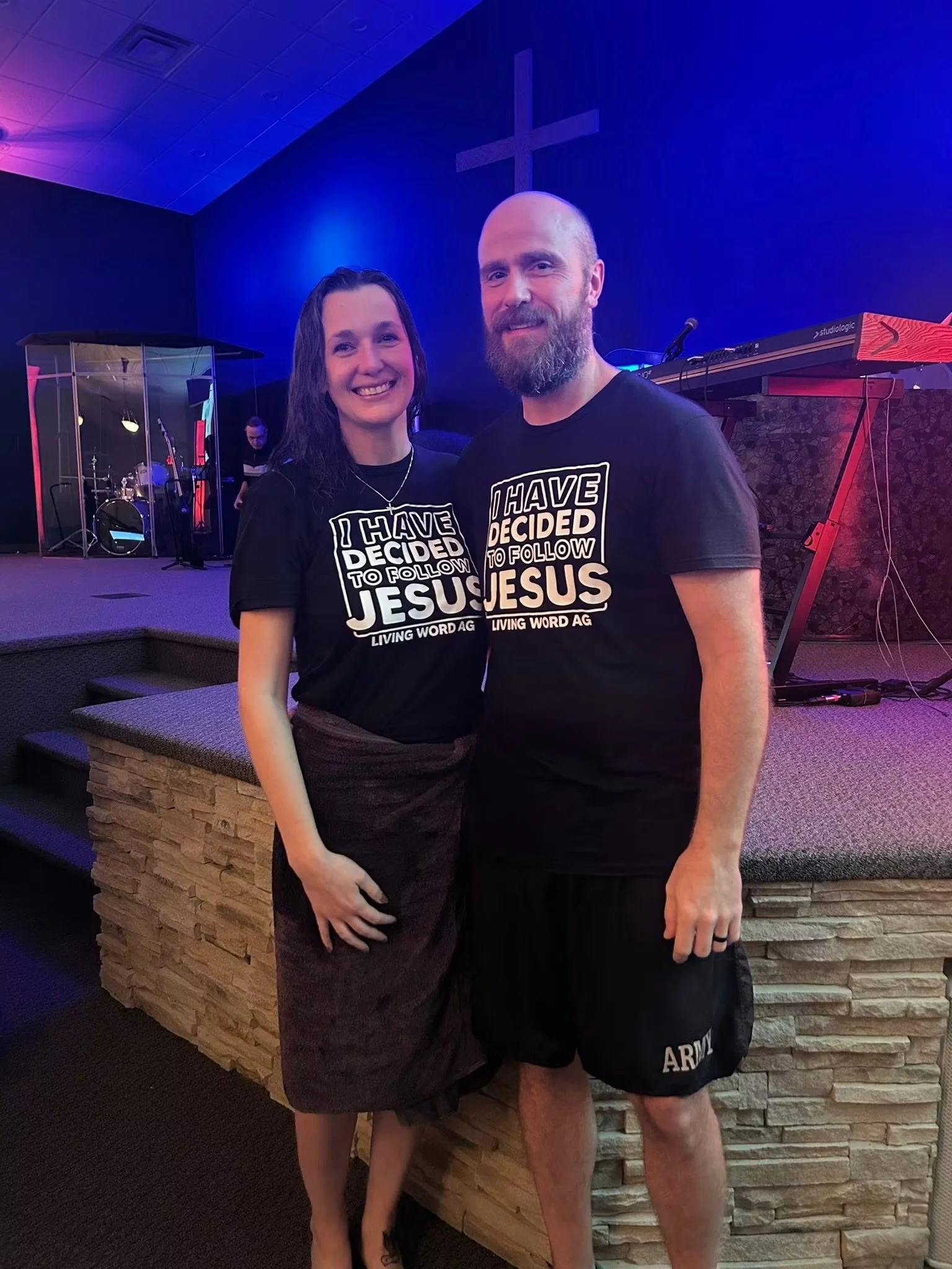 A man and woman standing together in a church interior, both smiling. They are wearing black T-shirts with white text that says, 'I HAVE DECIDED TO FOLLOW JESUS.' The woman has long dark hair and is wearing a brown skirt, while the man has a beard and is wearing black shorts. There is a wooden stage and musical instruments in the background, with purple and blue lighting and a cross on the wall.