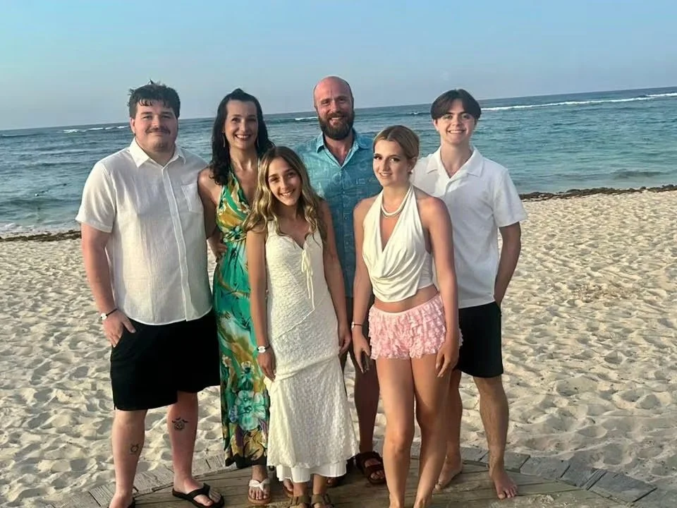 A group of six people, including men and women, standing on a sandy beach with the ocean in the background, dressed in casual summer clothing.