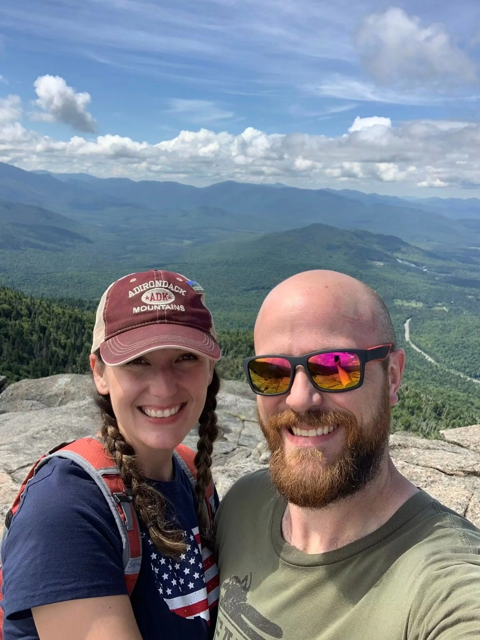 A man and a woman taking a selfie with a mountain view in the background. The woman is wearing a baseball cap and a shirt with an American flag pattern. The man has a shaved head, a beard, and sunglasses that reflect the sky.