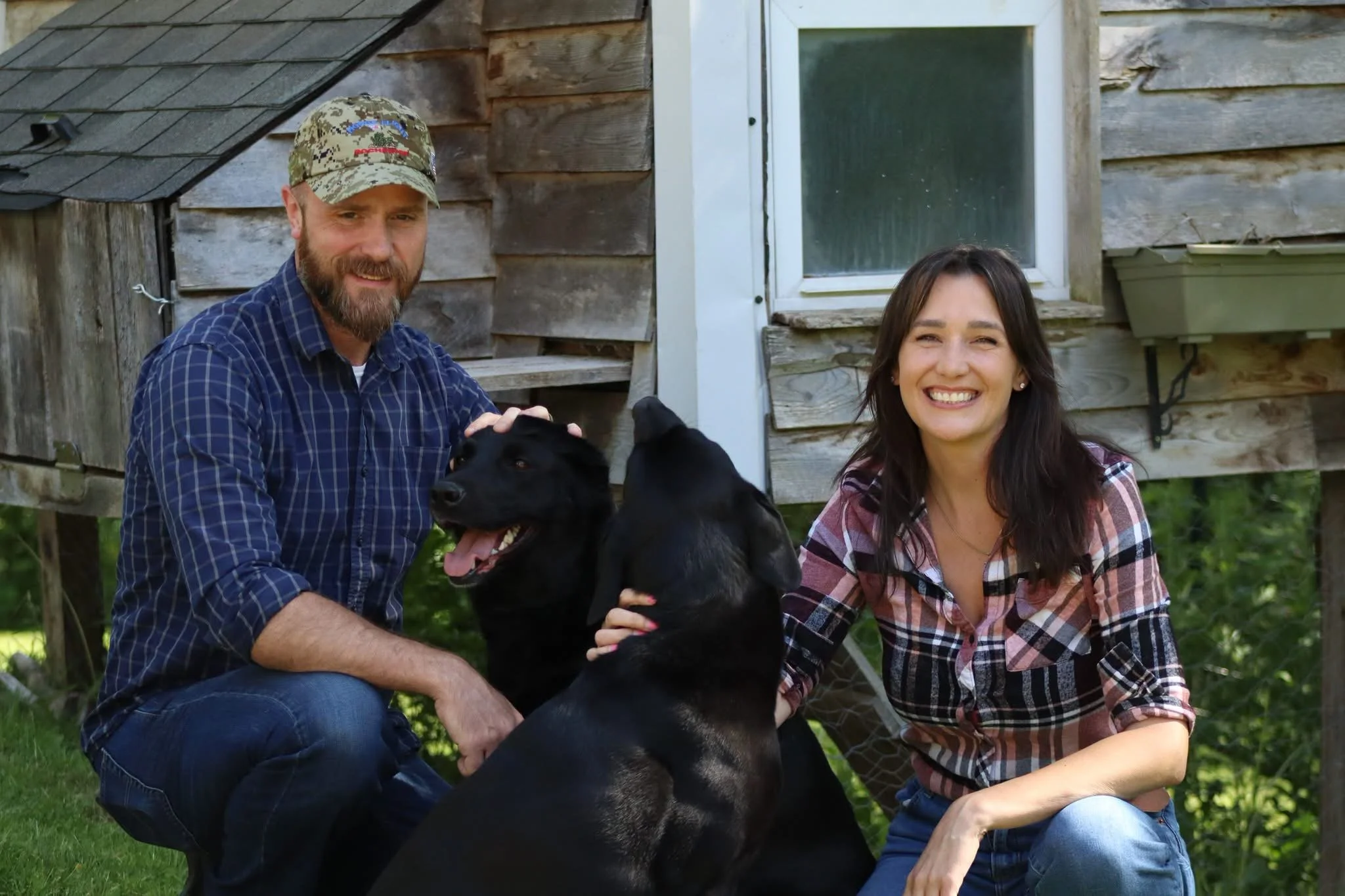 A man with a beard and baseball cap, and a woman with long dark hair, sitting outdoors in front of a rustic wooden building, smiling and holding a large black dog.