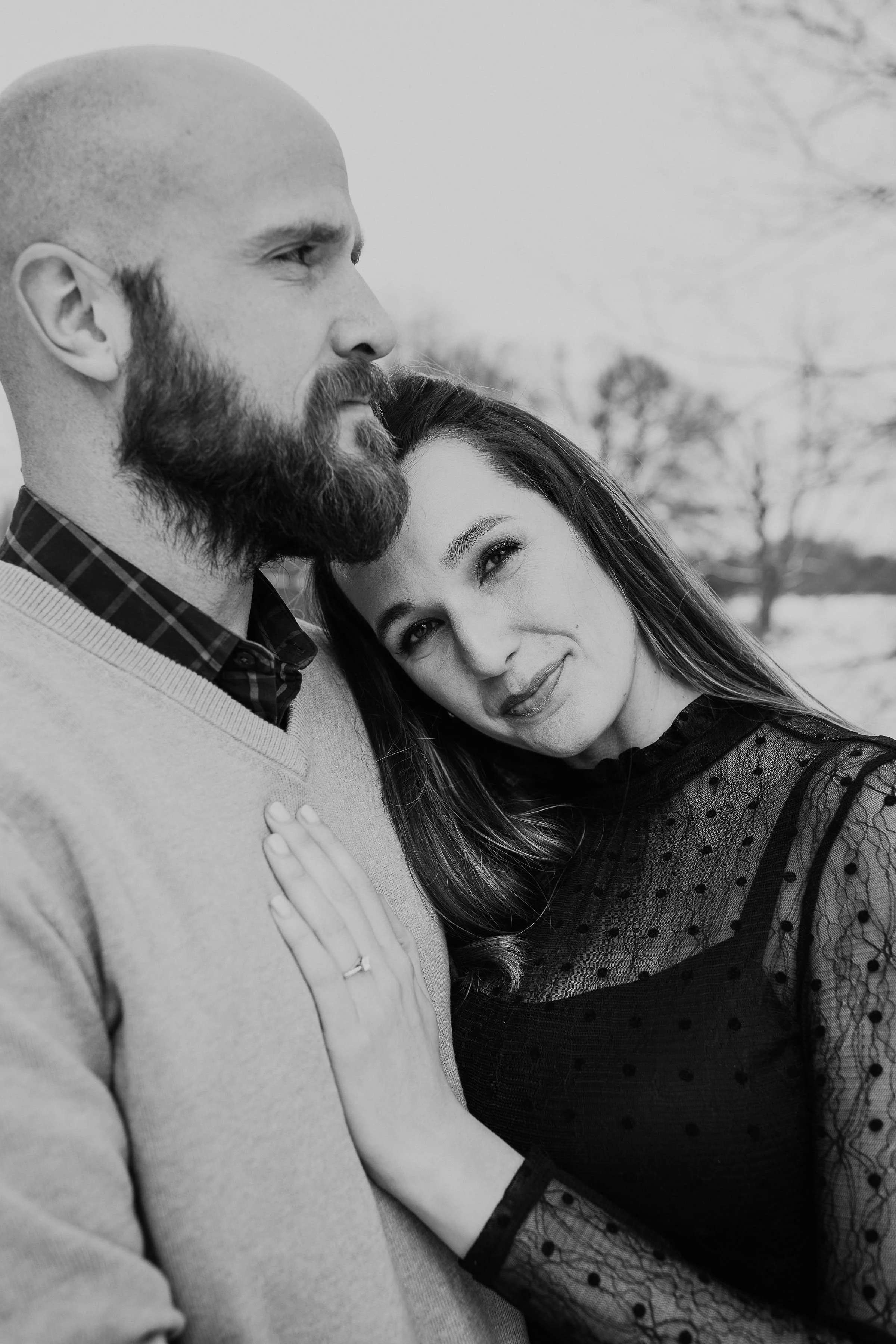 A black and white photo of a couple outdoors, with the woman leaning her head on the man's shoulder, both showing gentle expressions.