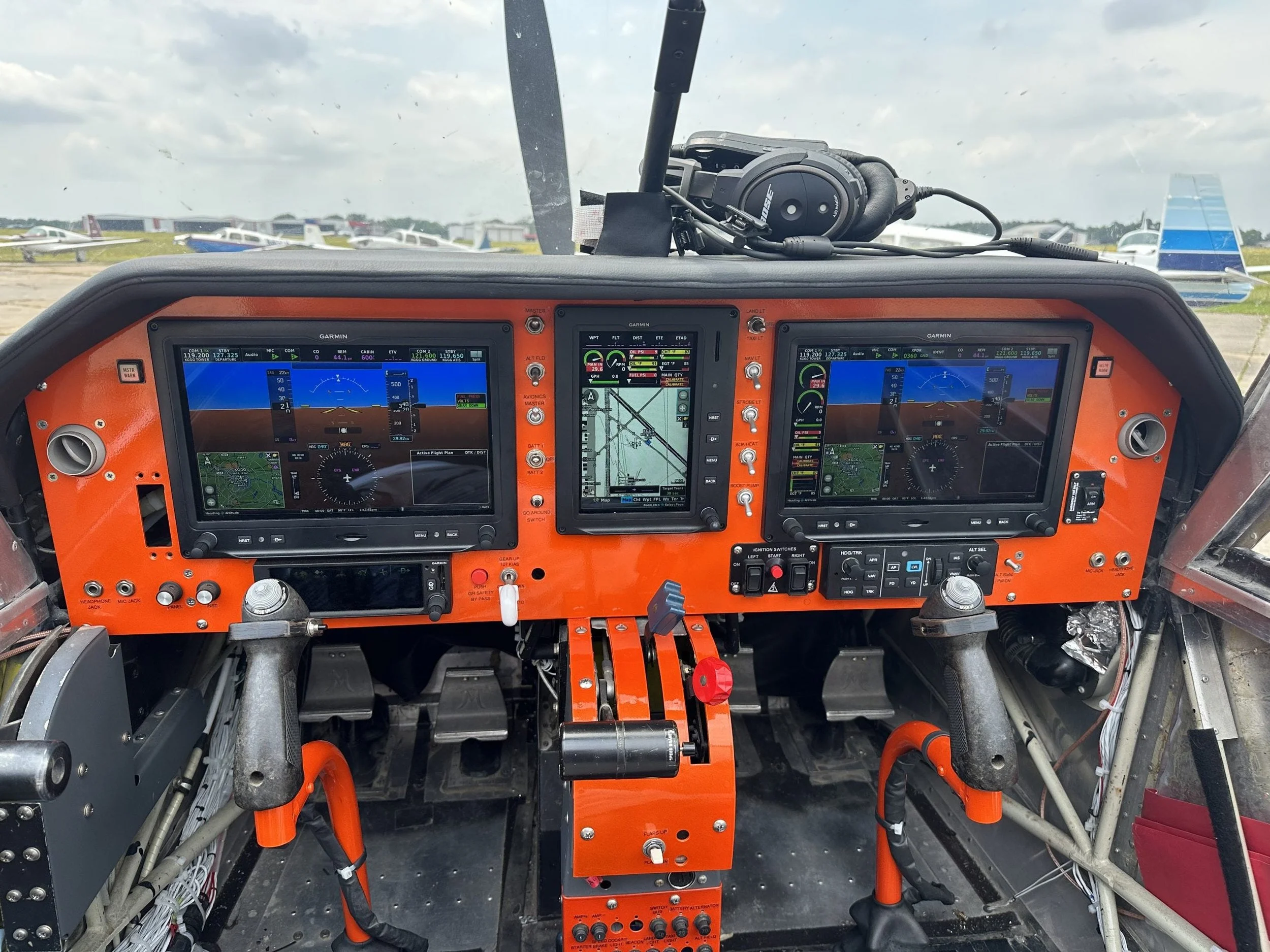 Interior of an aircraft cockpit with multiple digital displays, controls, and wiring. Outside the cockpit window, several small airplane aircrafts are parked on the tarmac under a cloudy sky.