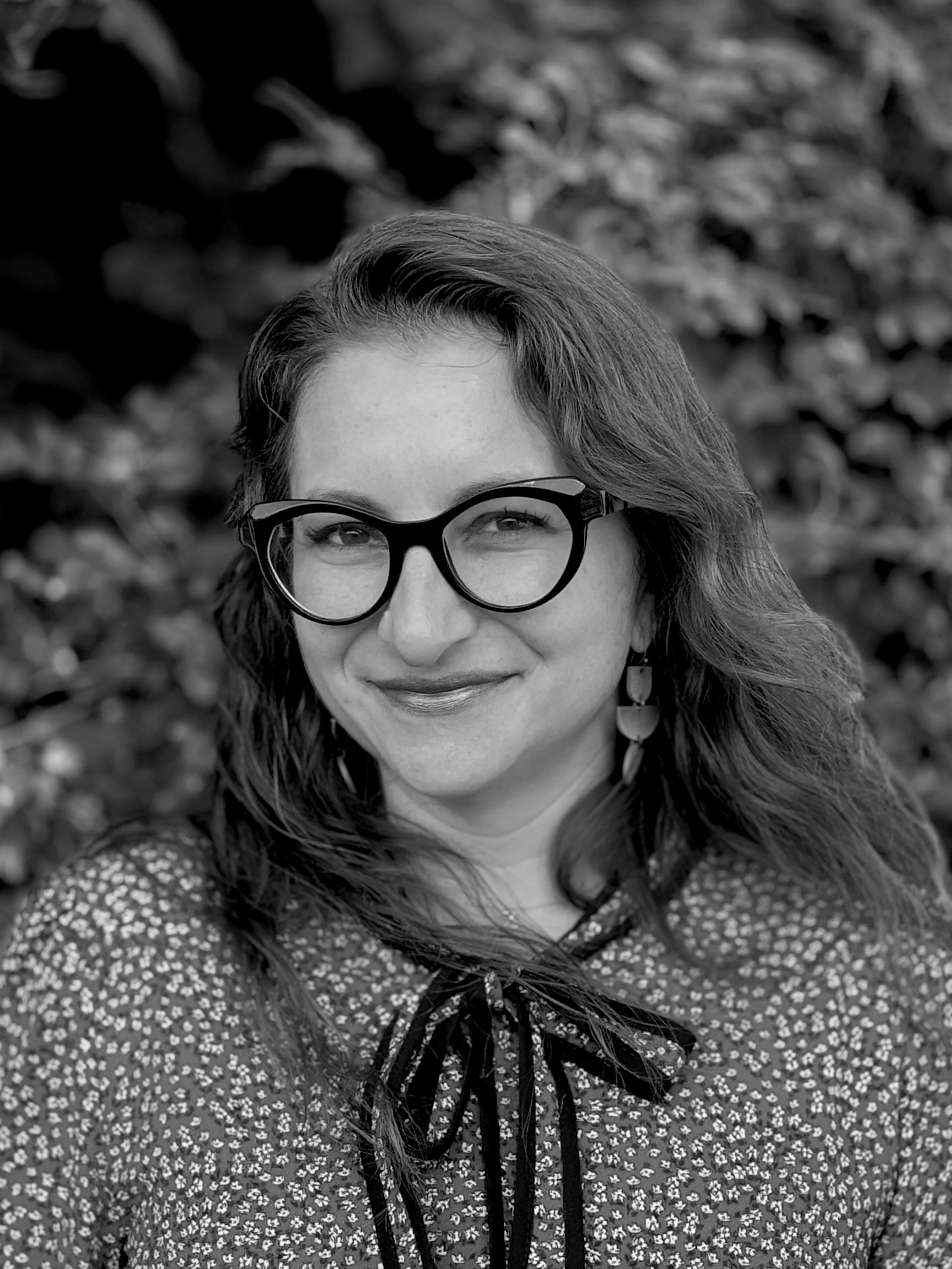 A black-and-white photograph of a woman with long wavy hair wearing glasses and earrings, smiling, with a blurred natural background.