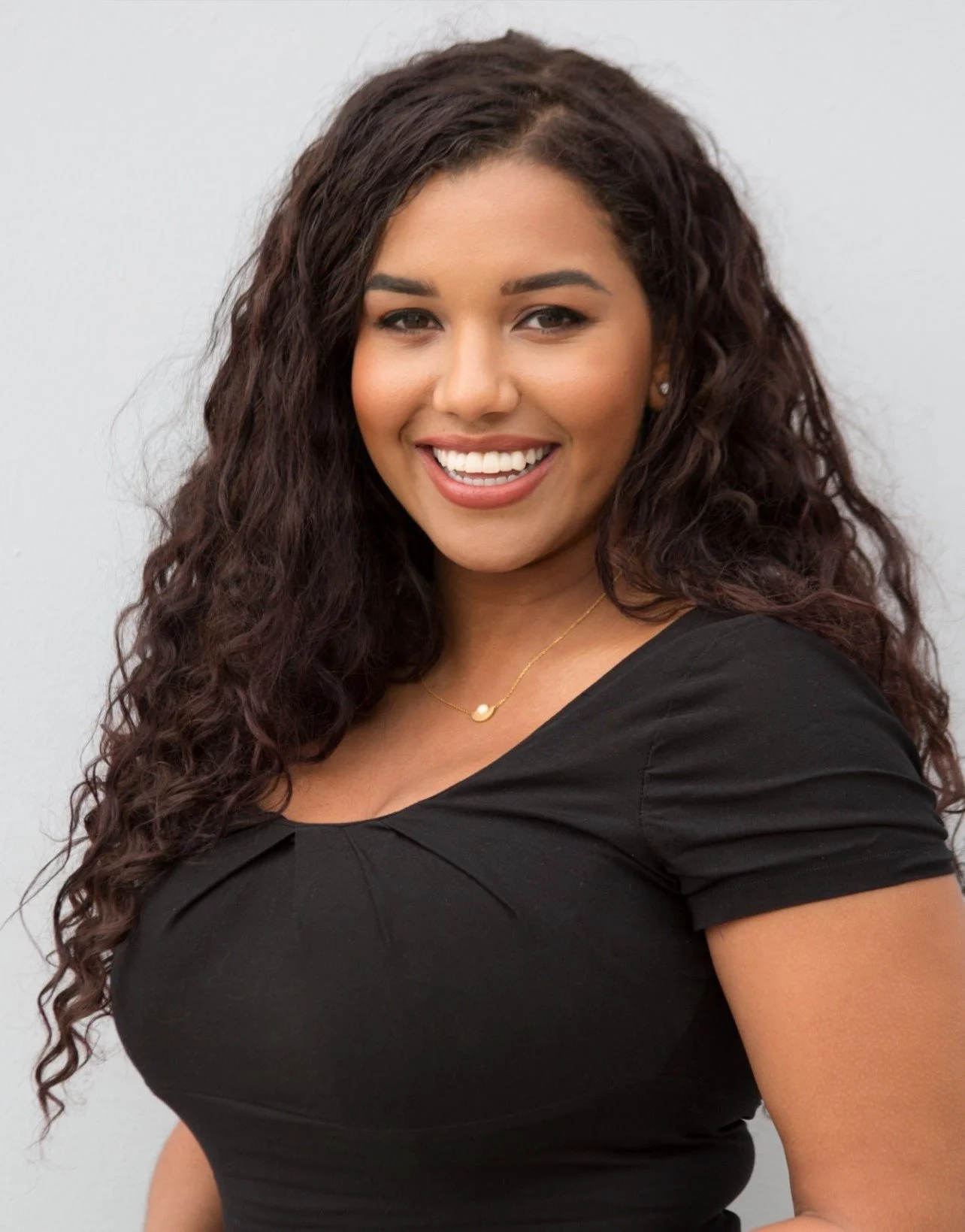 A woman with long, curly dark hair smiling, wearing a black top, a delicate gold necklace with a small pendant, and earrings, standing against a plain light gray background.