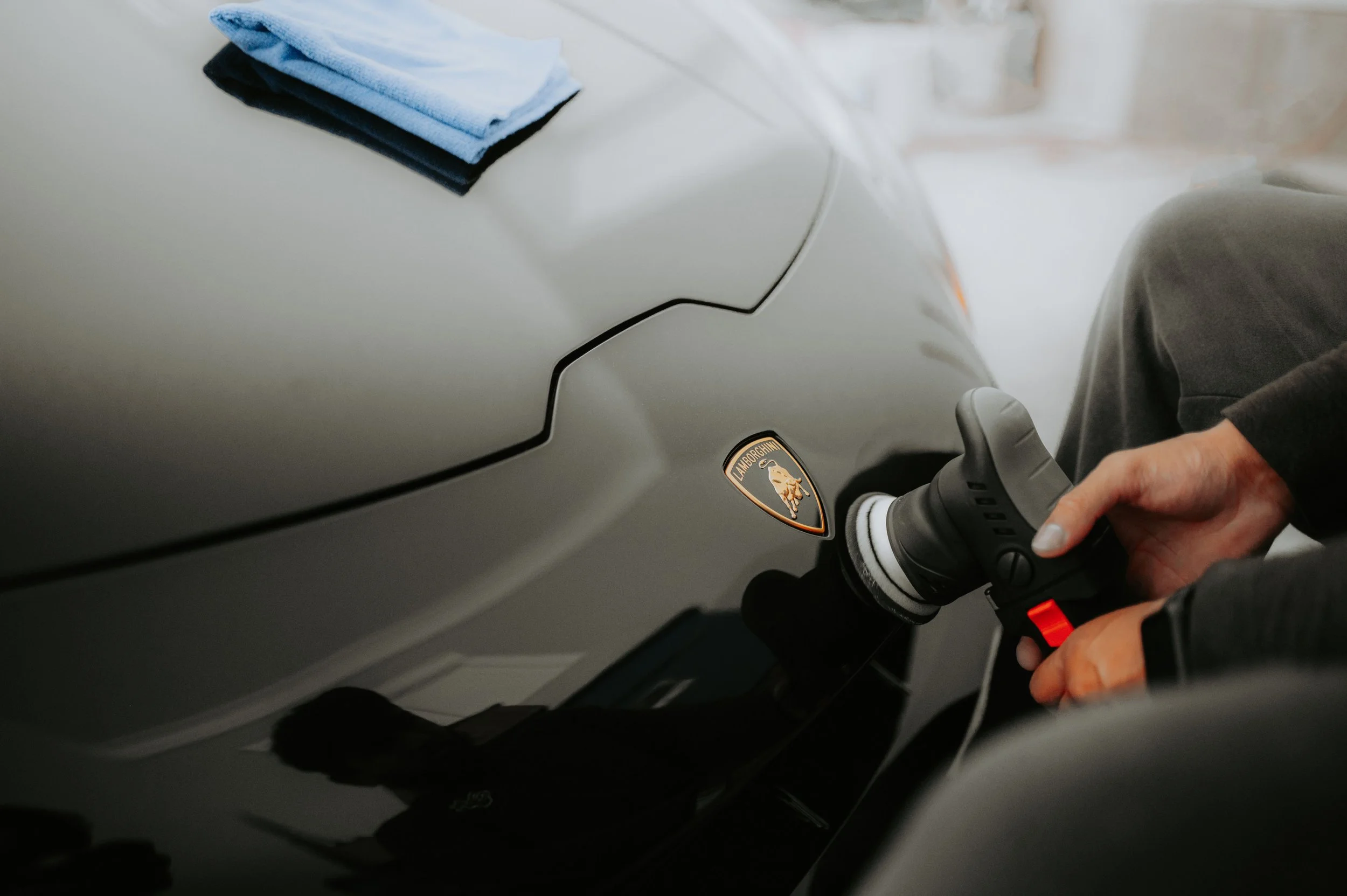 Person cleaning a grey Lamborghini with a handheld polisher.