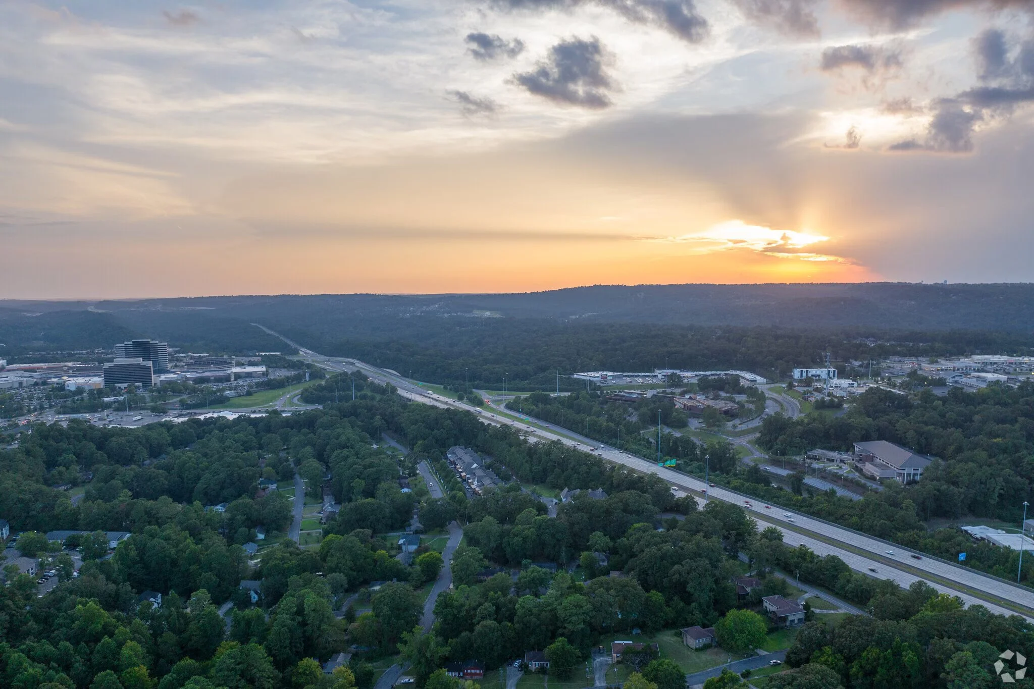 A cityscape at sunset with a mix of commercial and residential buildings, highways, and a lush green forest in the foreground. The sky has scattered clouds and the sun is partially hidden behind them, casting a warm glow over the scene.