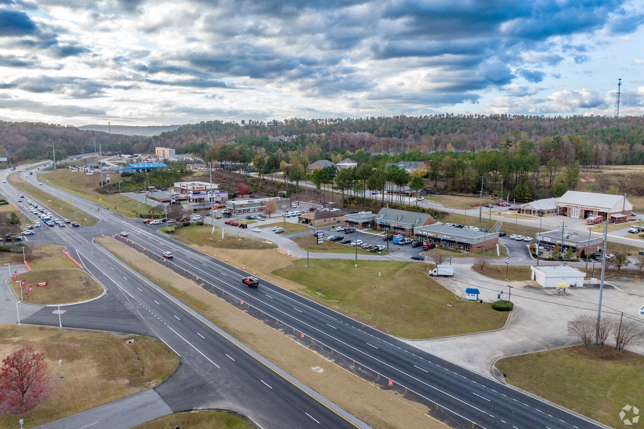 Aerial view of a commercial area with roads, parking lots, a McDonald's restaurant, and surrounding buildings, with a wooded hillside in the background and a cloudy sky overhead.