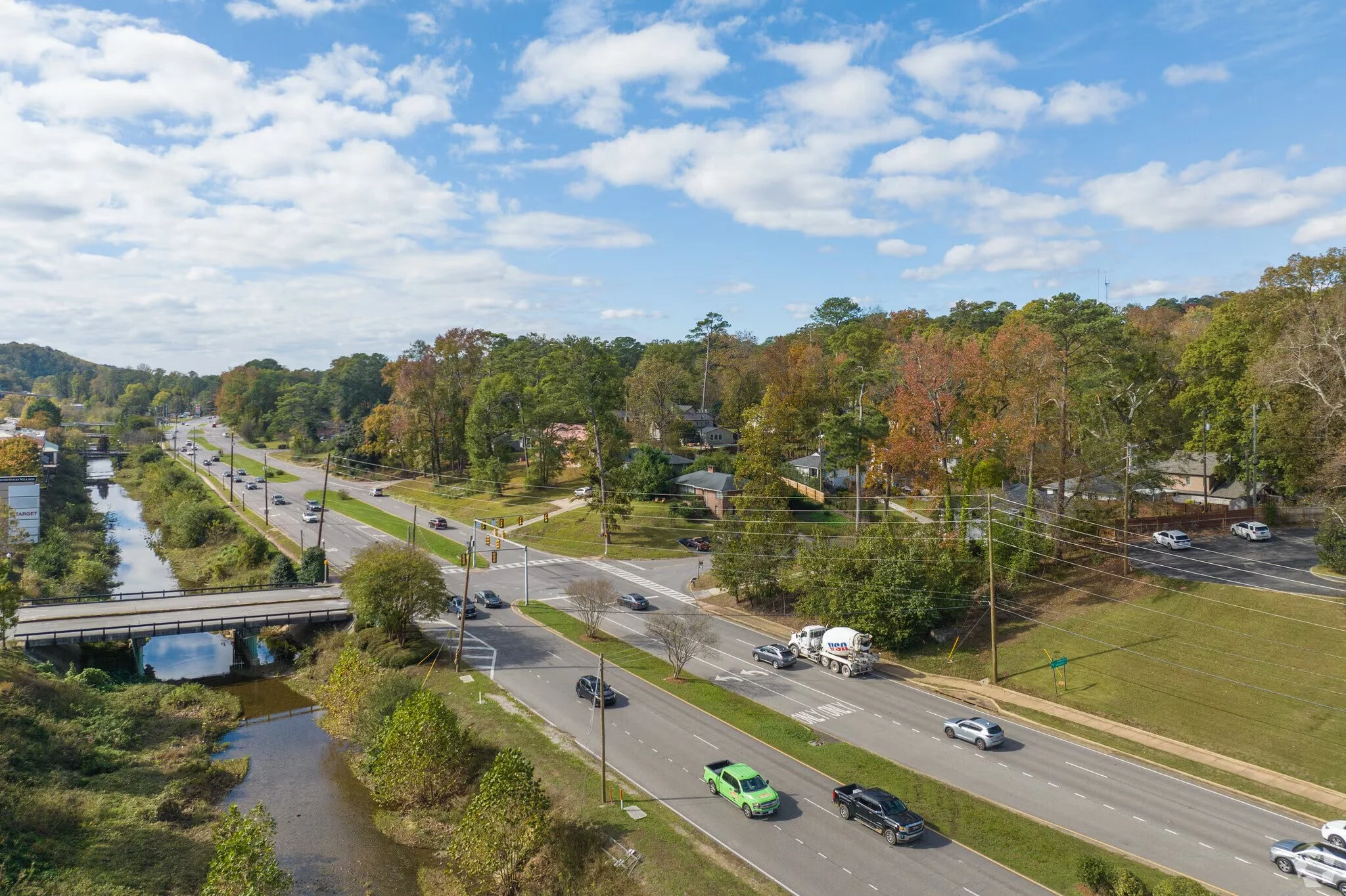 Aerial view of a small town with a river running parallel to a three-lane road. Traffic, trees with autumn foliage, houses, and a bridge over the river are visible under a partly cloudy sky.