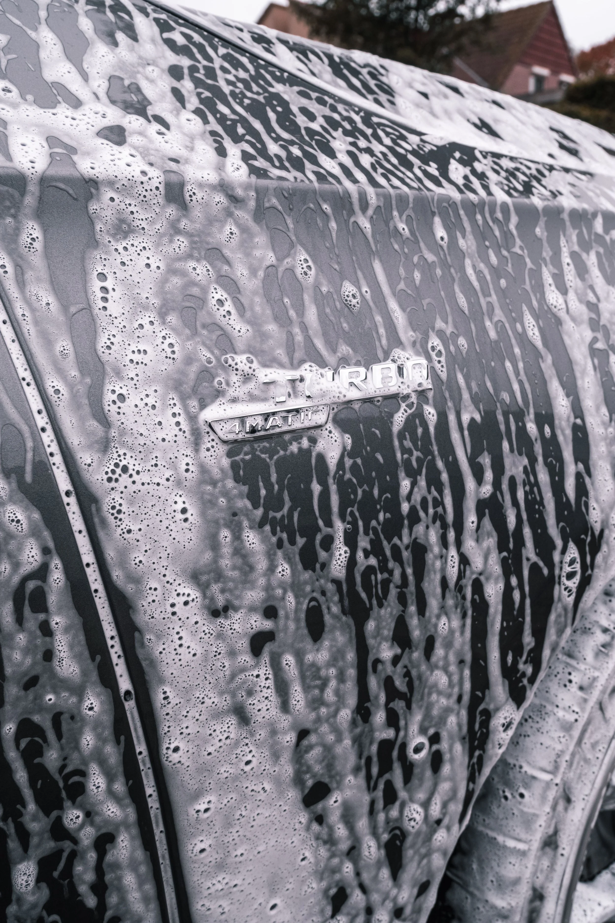A close-up of a silver car being washed, covered with soap suds and foam.