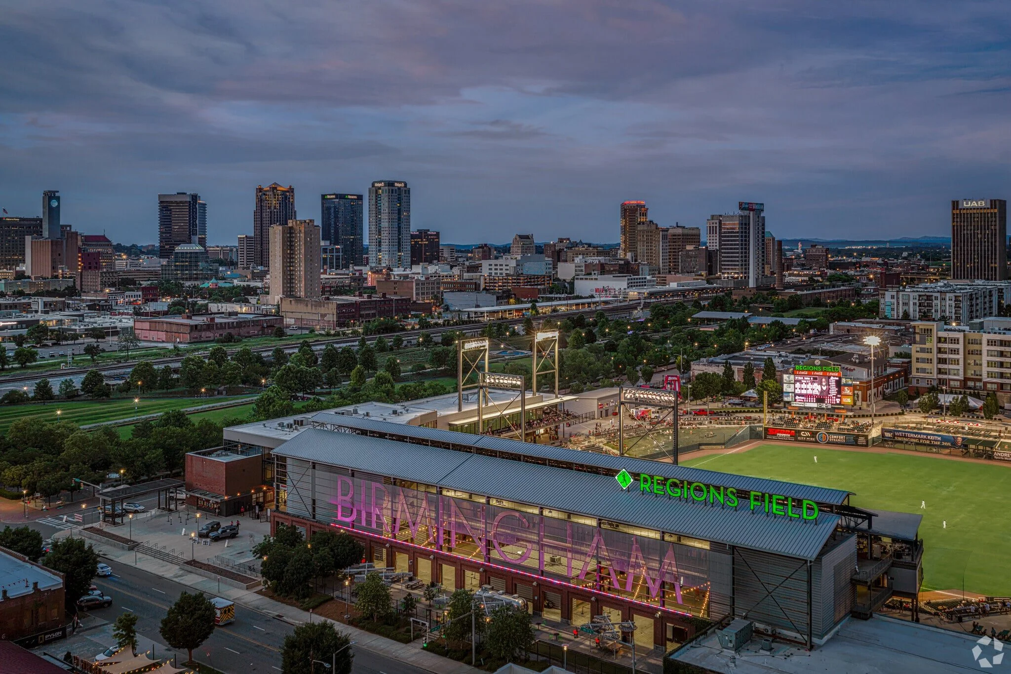 A city skyline at dusk with tall buildings, some with illuminated signs, and a baseball stadium called 'Regions Field' in the foreground. The stadium has a sign with pink and green lighting, a field with players, and surrounding parking lots and streets.