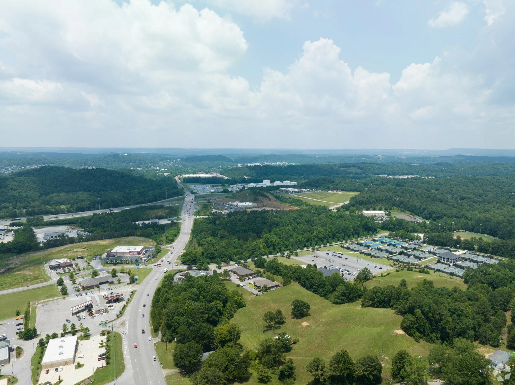 Aerial view of a landscape with a highway, commercial buildings, trees, grassy areas, and a cloudy sky overhead.