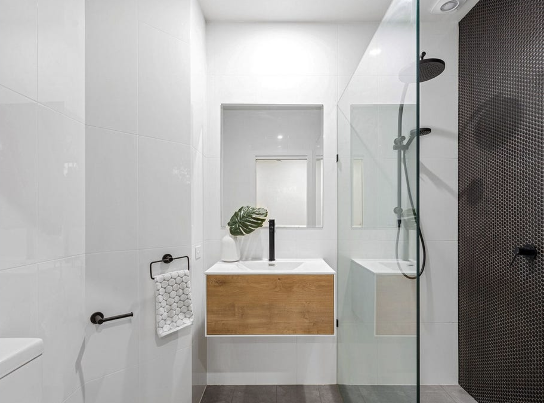 Modern bathroom with white tiled walls, a black showerhead and a glass partition, a wooden vanity with a white sink, a large green leaf in a white vase, a small black soap dispenser, and a white towel on a black towel holder.