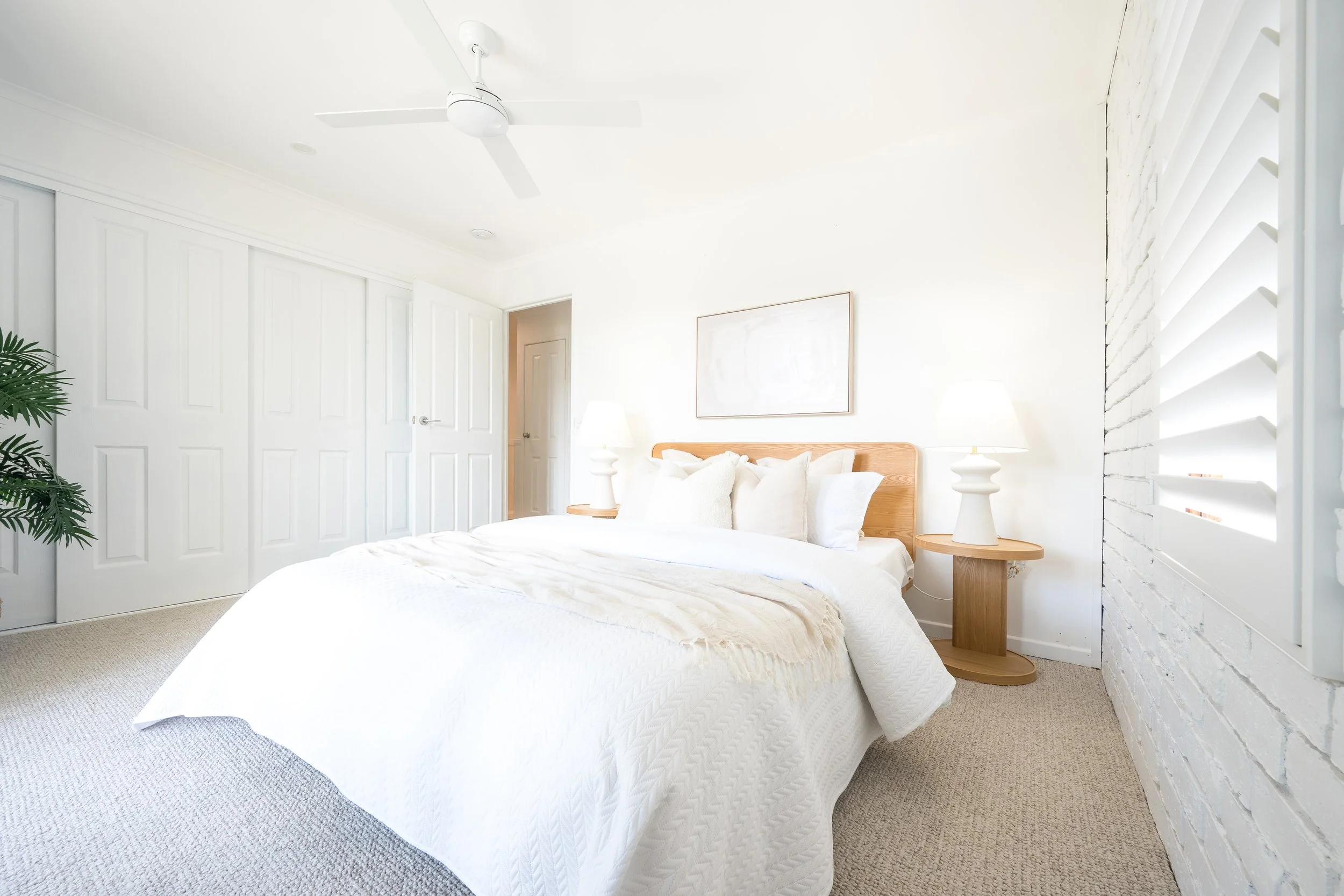 Bright, white bedroom with a bed, nightstands, lamps, and a ceiling fan. Features a white brick wall and louvered shutters.
