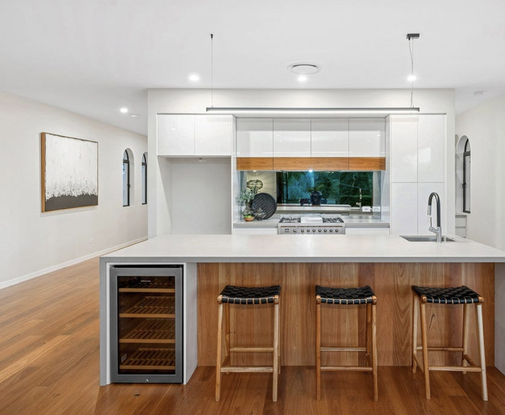Modern kitchen with white cabinets, wooden accents, a large white island with three black stools, and a window behind the stove showing trees outside.