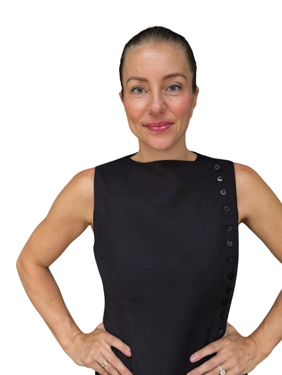 A woman with light skin, brown hair tied back, wearing a sleeveless black dress with buttons along the shoulder and side, smiling with her hands on her hips against a white background.