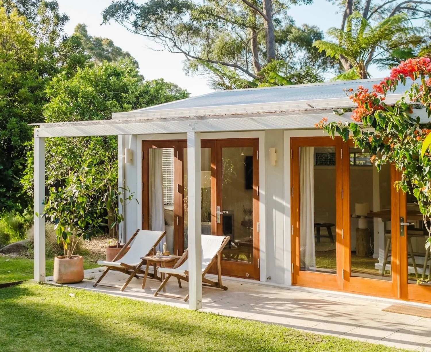 Small modern house with wooden framed glass doors and a white metal roof, surrounded by green trees and a lawn, with two white deck chairs and potted plants on a patio