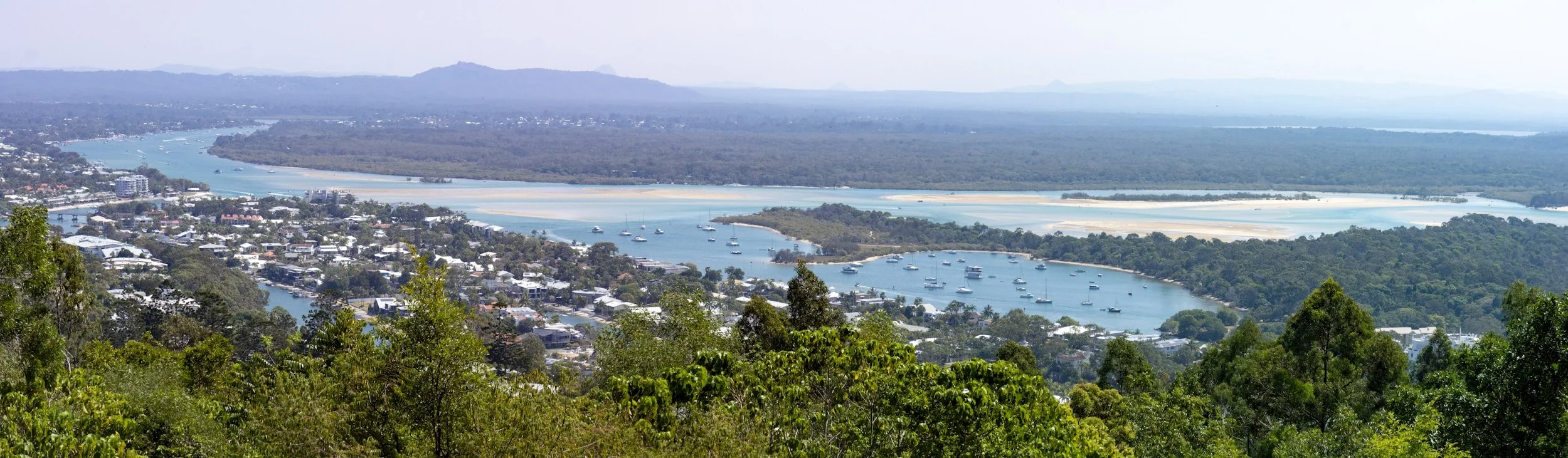 A panoramic view of a river with boats, surrounded by a town with buildings, and dense green forest in the foreground and distant hills in the background.