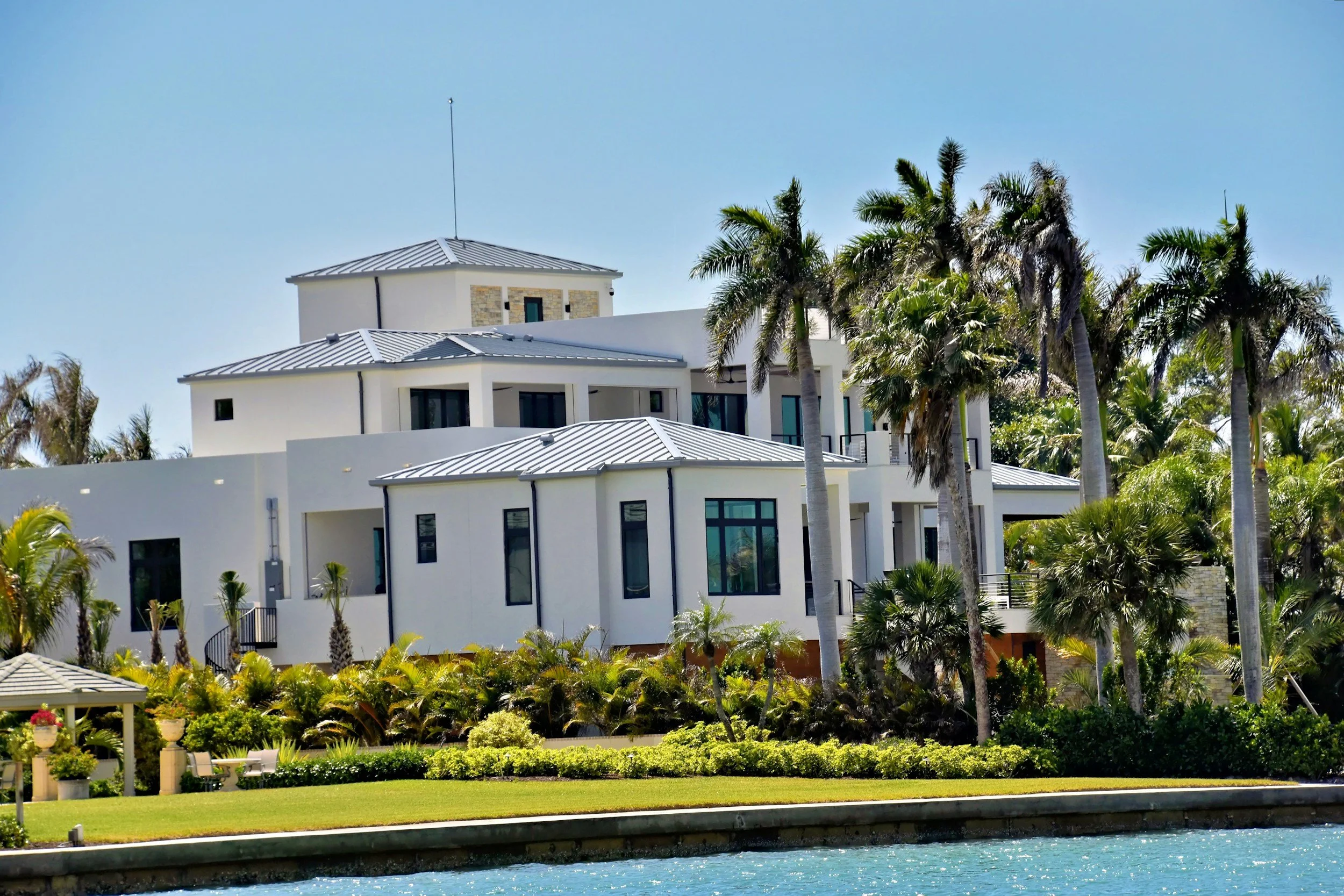 Luxury modern white house with multiple levels and gray metal roofs, surrounded by tall palm trees and lush landscaping, next to a waterway under a clear blue sky.