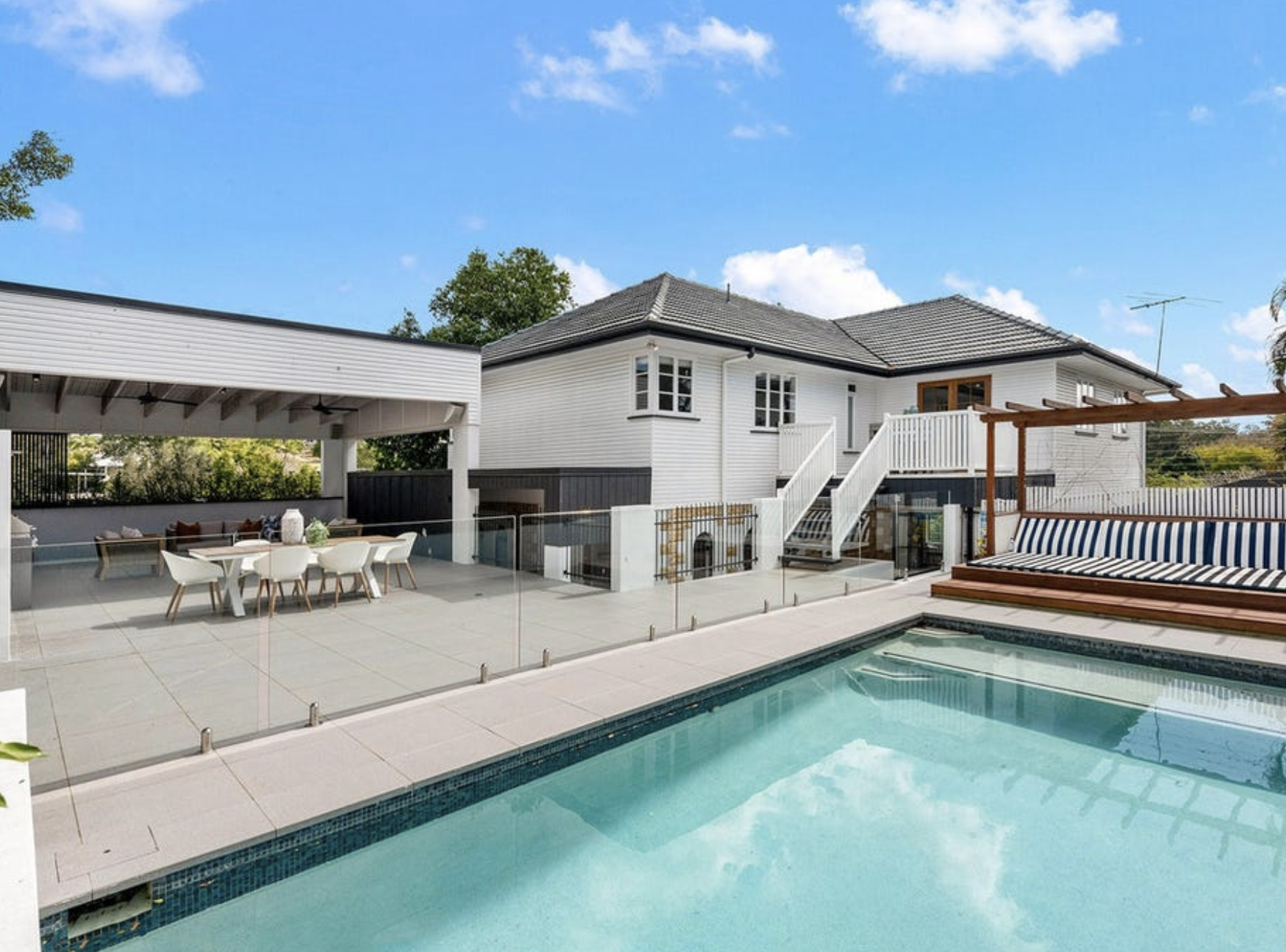 Modern backyard with swimming pool, outdoor dining area with white chairs and table, lounge with striped cushion, and a white house with stairs and a balcony under a blue sky with scattered clouds.