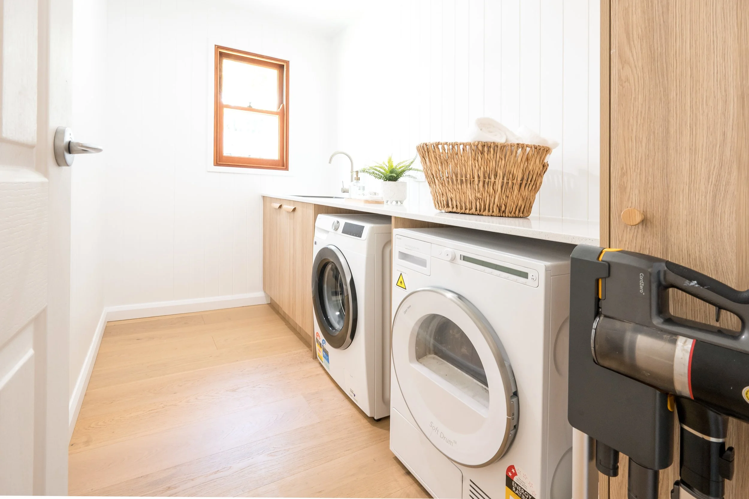 A laundry room with a wooden floor, white walls, a small window with wooden trim, a countertop with a plant and laundry supplies, a washing machine, an electric dryer, a wicker basket with folded towels, and cleaning tools.