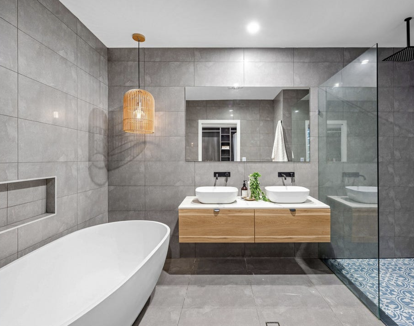 Modern gray-tiled bathroom with freestanding bathtub, double vanity with vessel sinks, large mirror, pendant light, and walk-in shower with glass partition.