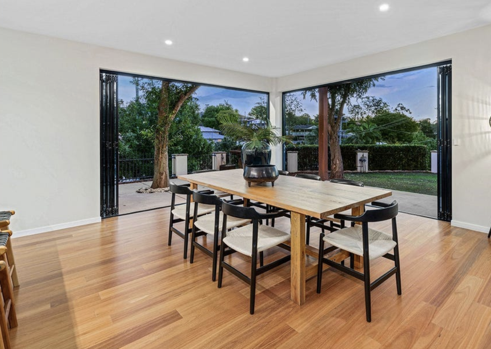 Dining room with wooden floor, rectangular table, six chairs, large windows showing outdoor trees and yard.