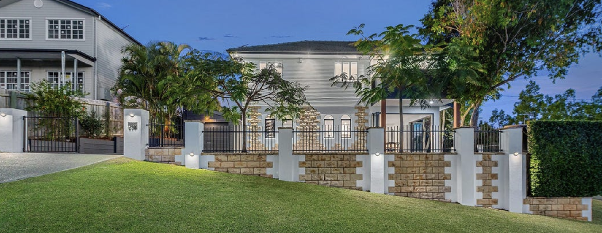 A modern house with a white exterior, stone accents, and a front yard decorated with green grass and trees. The house features large windows, a balcony, and a black fence surrounding the yard, illuminated at dusk.