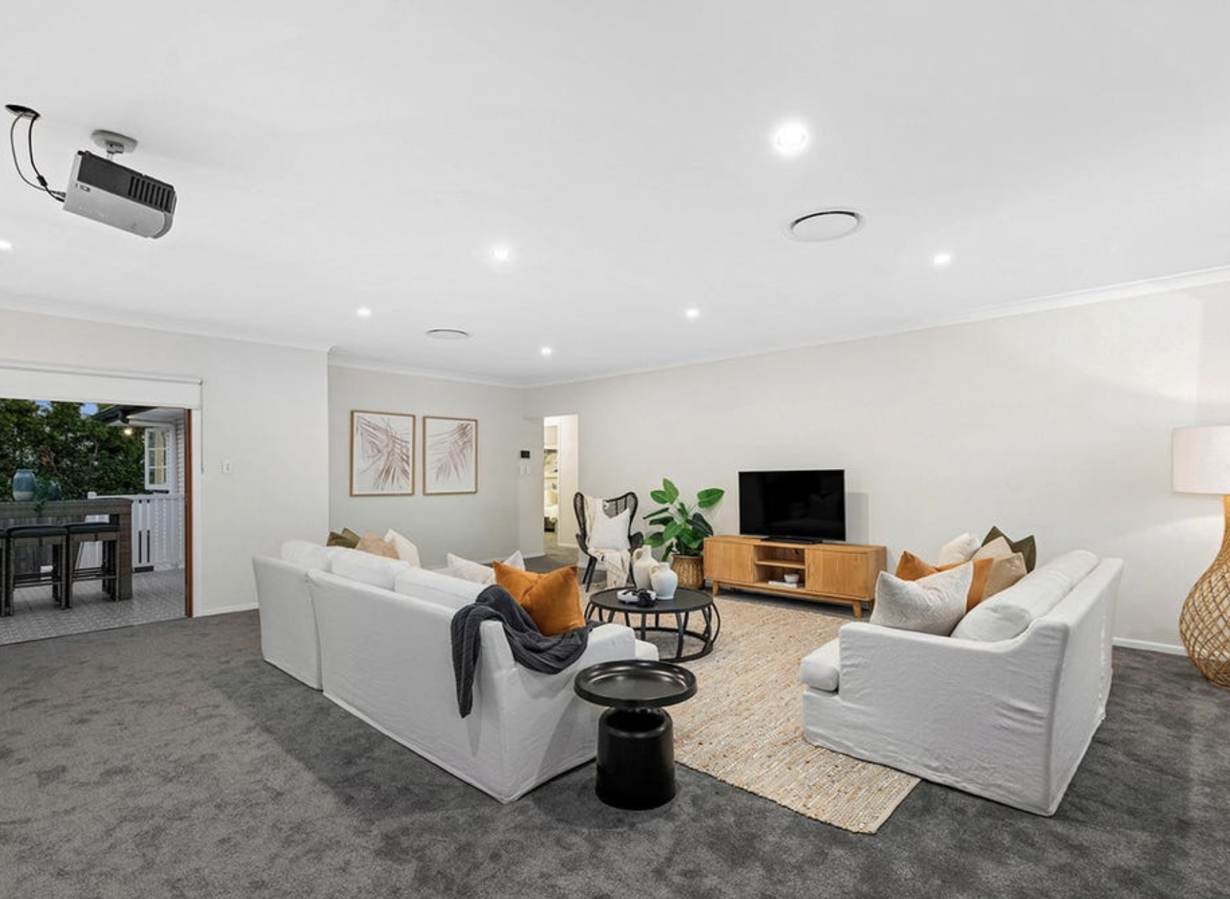Living room with white sofas, a black coffee table, a wooden TV stand with a flat-screen TV, decorative pillows, houseplants, and artwork on the walls.
