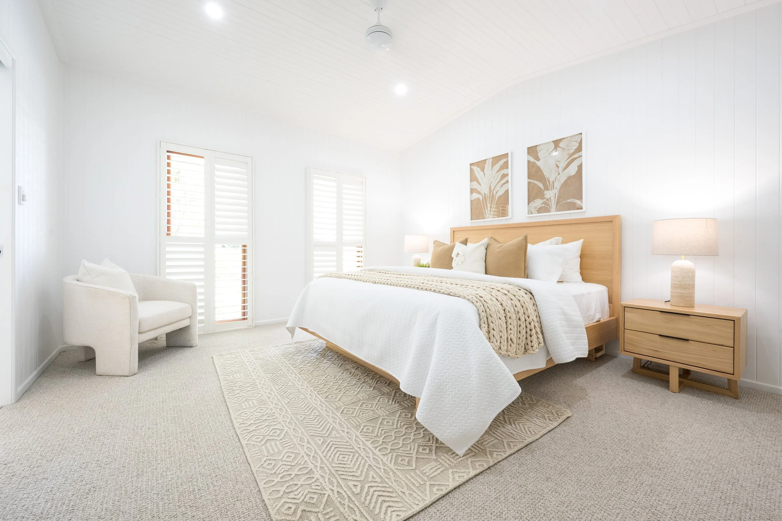 A bright, minimalist bedroom featuring a large bed with beige and white pillows and a knitted throw. There are two framed botanical prints above the bed, matching bedside tables with lamps, a plush armchair, and windows with white shutters. The room 