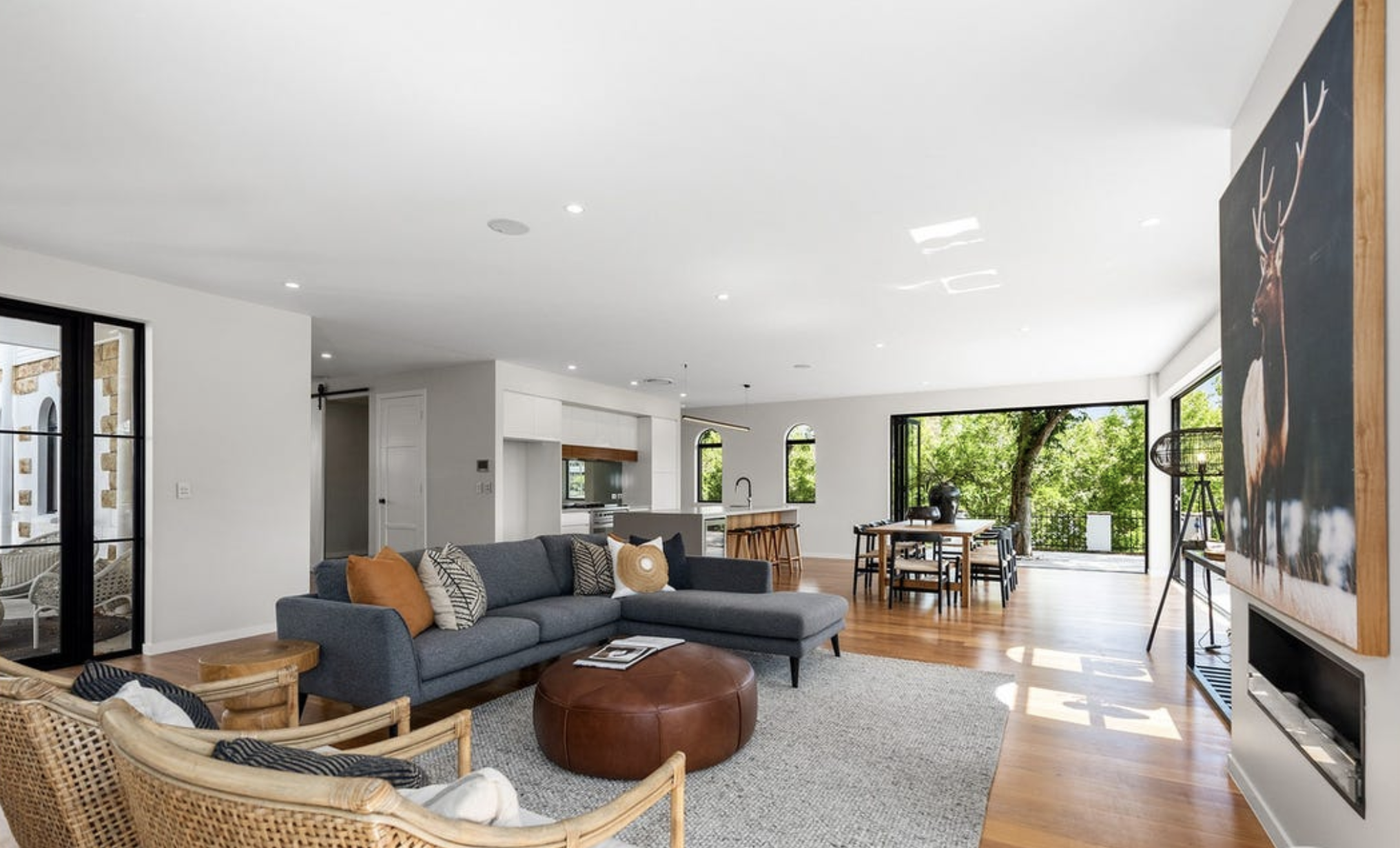 Modern open-concept living room with gray sofa, rattan chairs, orange pillows, large windows showing greenery outside, and a high ceiling with recessed lighting.