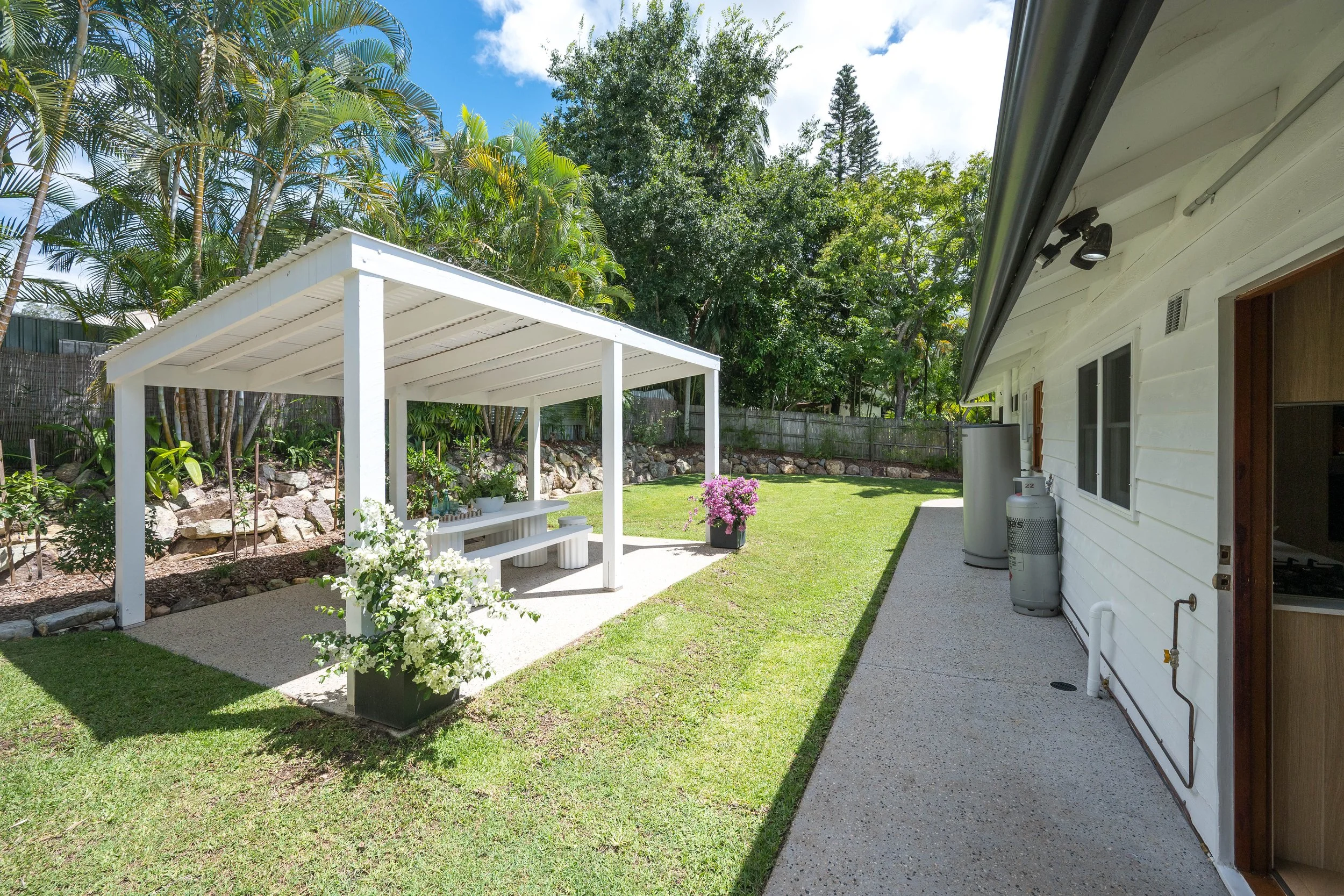 A backyard with a white covered patio, green grass, flowering plants, trees, and a white house with windows and an outdoor propane tank.