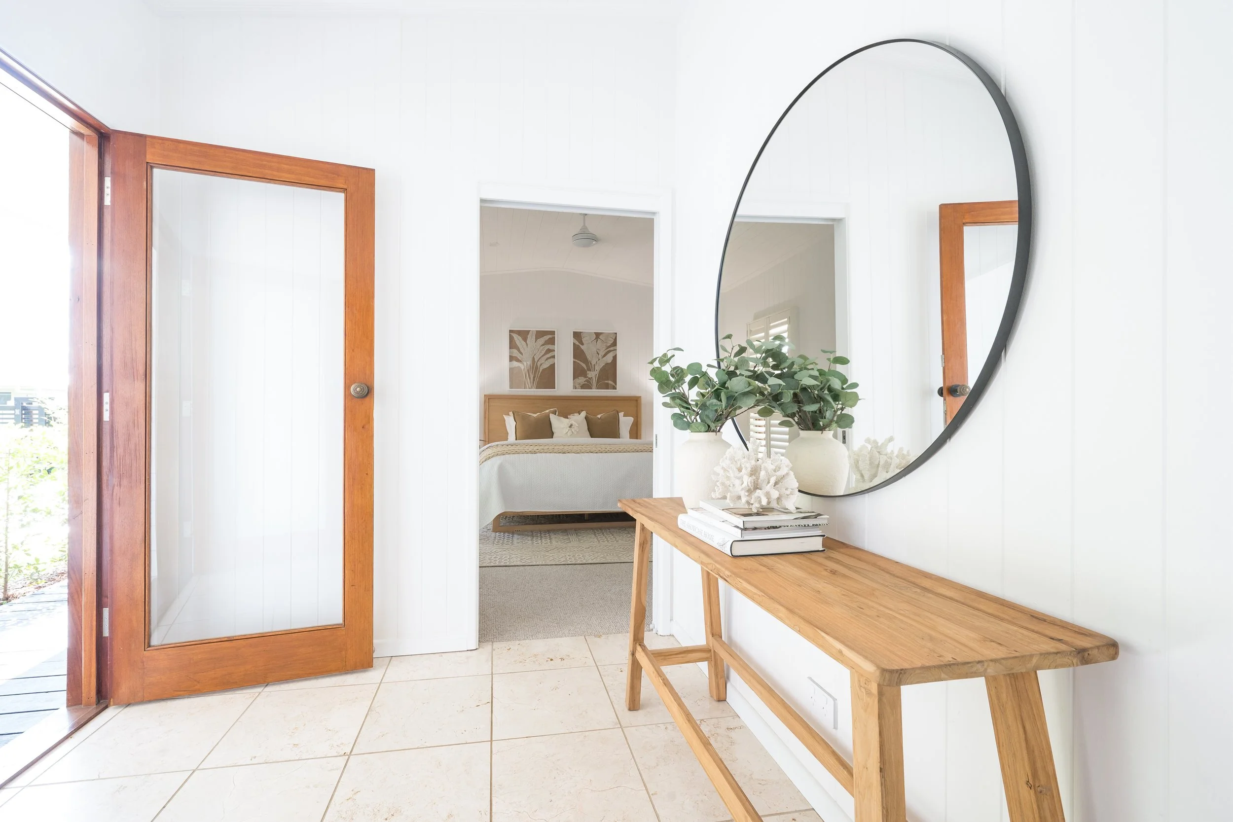 Entryway with a wooden door, a mirror on the wall, a wooden bench, and decorative plants on a side table leading into a bedroom with framed artwork and a bed.