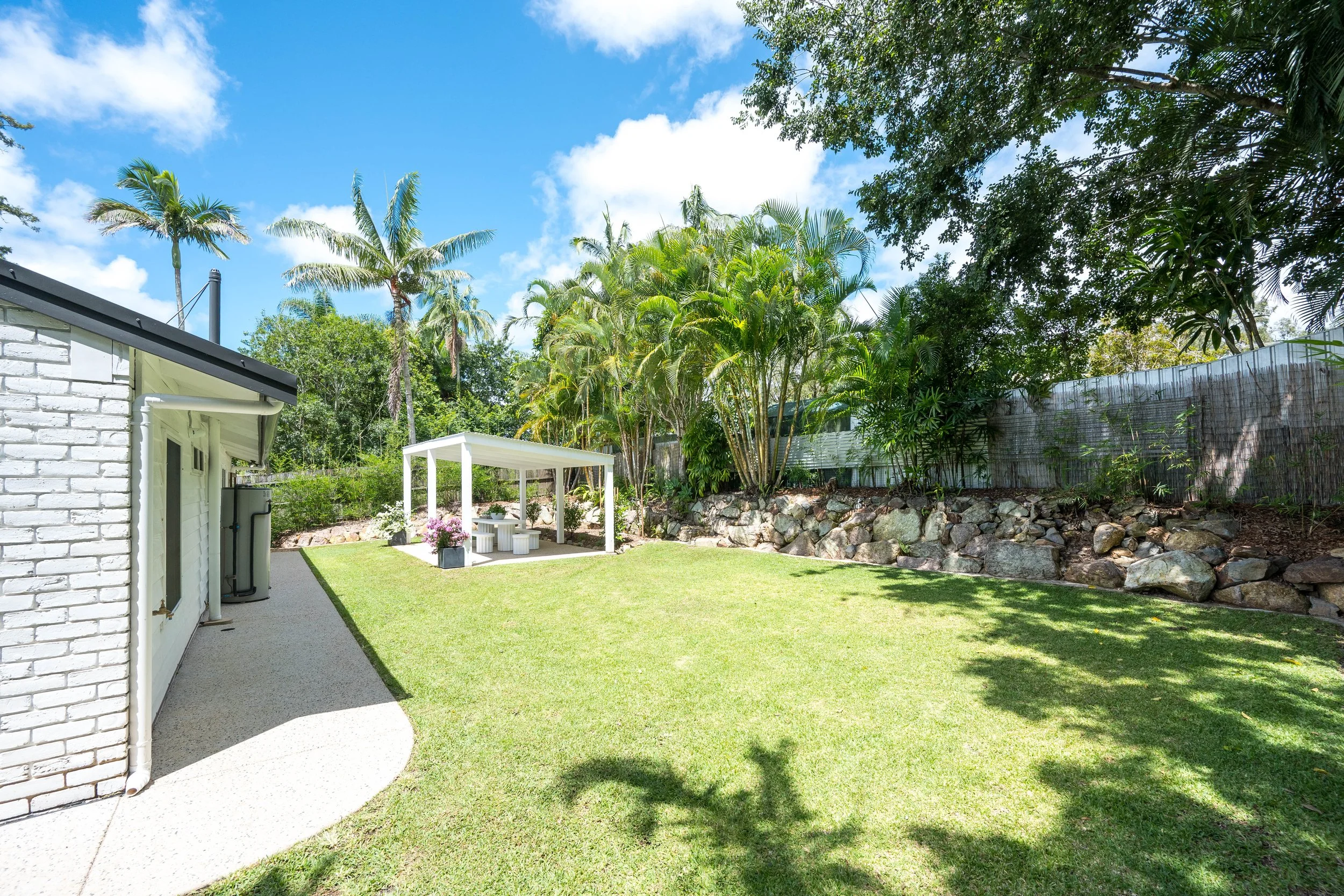 Backyard with green lawn, white patio table, and chairs in a shaded area, surrounded by trees, with a fence and a clear blue sky.