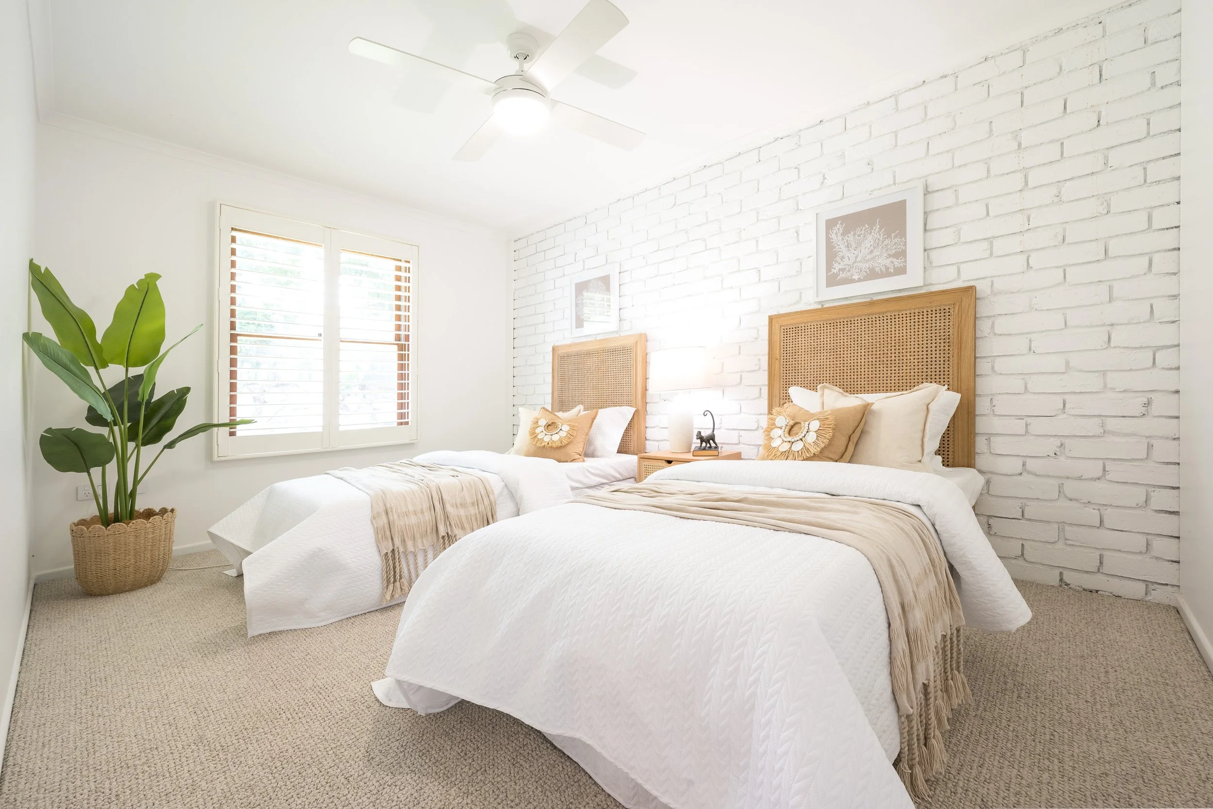 Bright bedroom with two beds that have white quilted covers and beige blankets, wooden headboards, tribal-pattern pillows, white walls with framed art, a white ceiling fan, a window with white blinds, a potted plant, and a nightstand with decor items