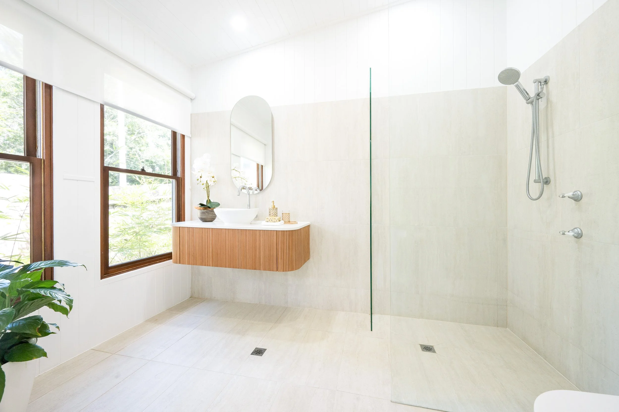 Modern bathroom with large windows, a walk-in shower with a glass divider, a white vessel sink on a wooden vanity, a mirror above the sink, some plants, and decorative items.