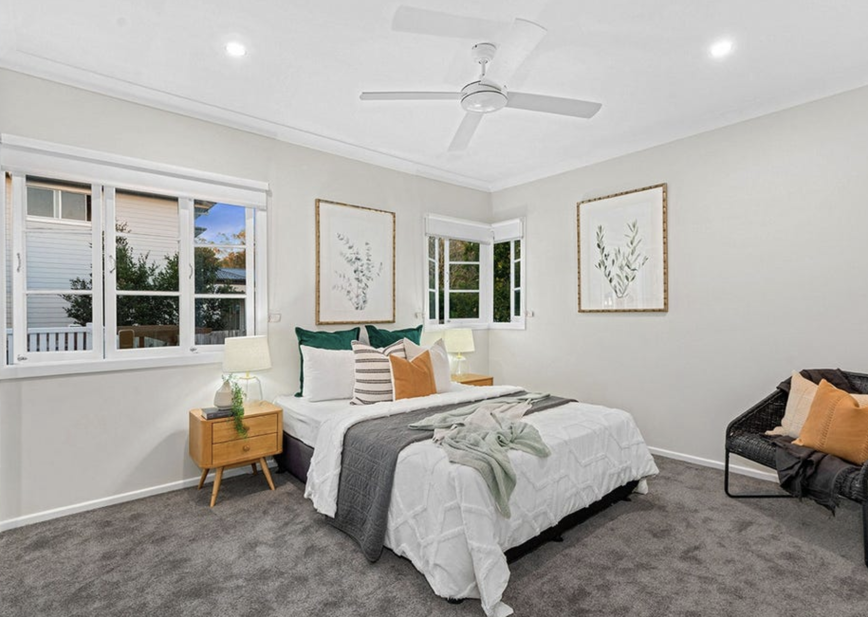 A cozy bedroom with a bed decorated with green, white, and orange pillows, a gray blanket, and a beige throw. There are two nightstands with lamps, framed botanical art on the walls, and a window showing trees outside. The room has a ceiling fan and 