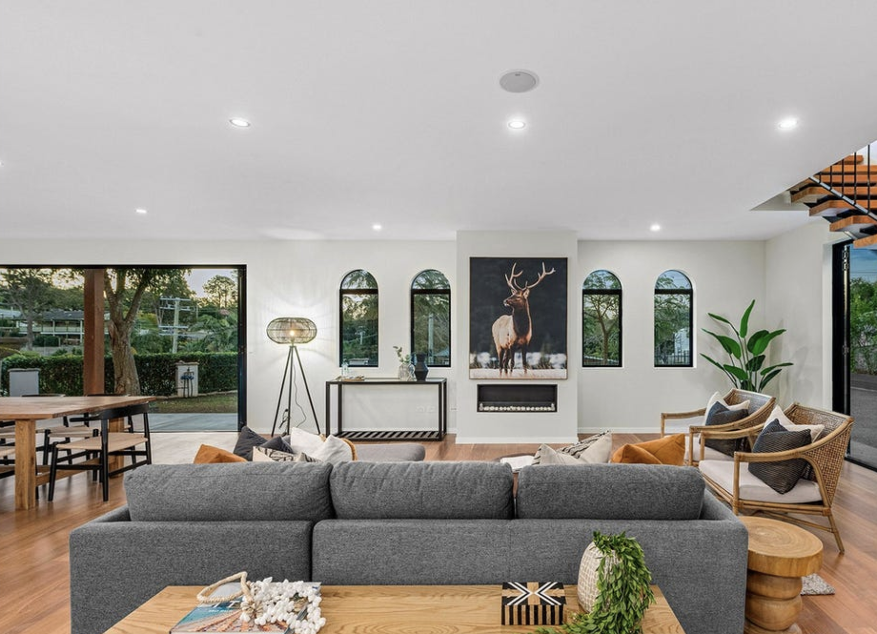Open living room with white walls, a large gray sofa, and wooden furniture, featuring a fireplace with a deer portrait above and large windows showing a garden outside.