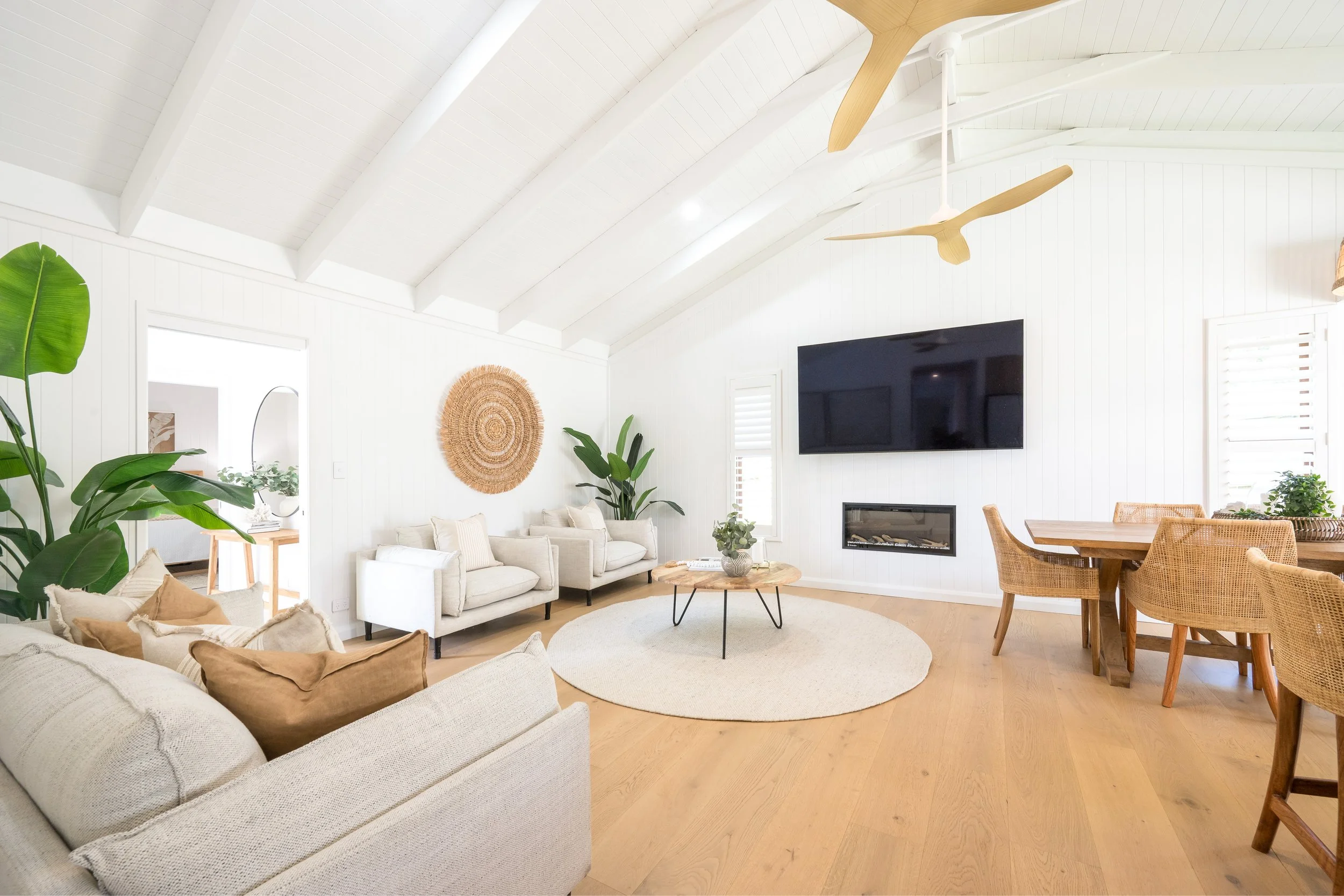 Bright living room with white walls, vaulted ceiling with exposed beams and ceiling fans, large television, sectional sofa, and wicker chairs, decorated with plants and woven wall art.