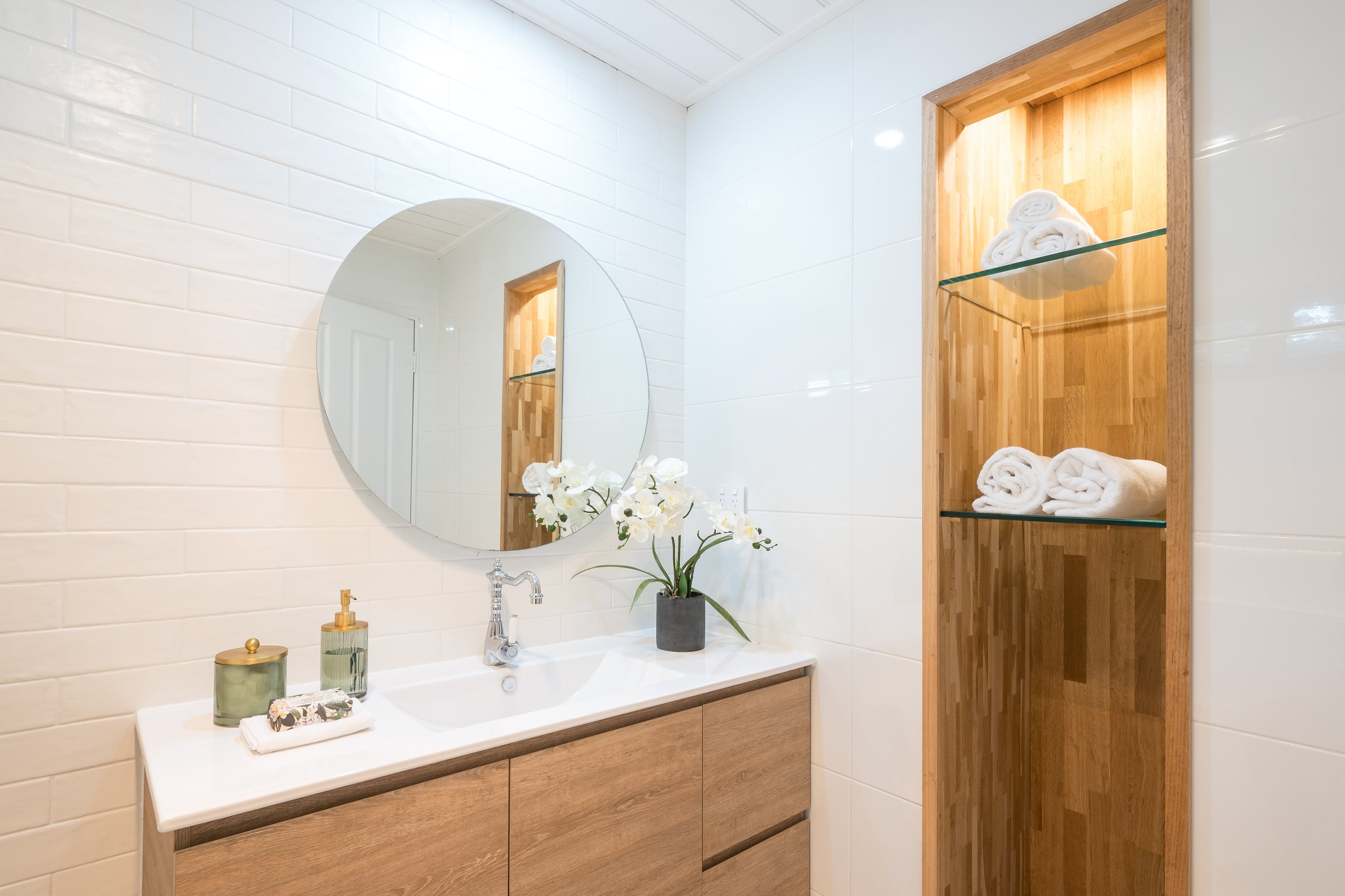 Bathroom vanity with a round mirror, a white sink, and a wooden cabinet with folded white towels on glass shelves, decorated with white flowers in a gray pot, and two green containers on the countertop.