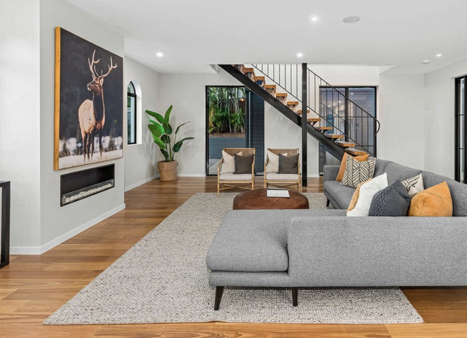 Modern living room with gray sectional sofa, decorative pillows, wooden coffee table, two chairs, fireplace, large artwork of a deer, potted plant, staircase, and sliding glass door leading outside.