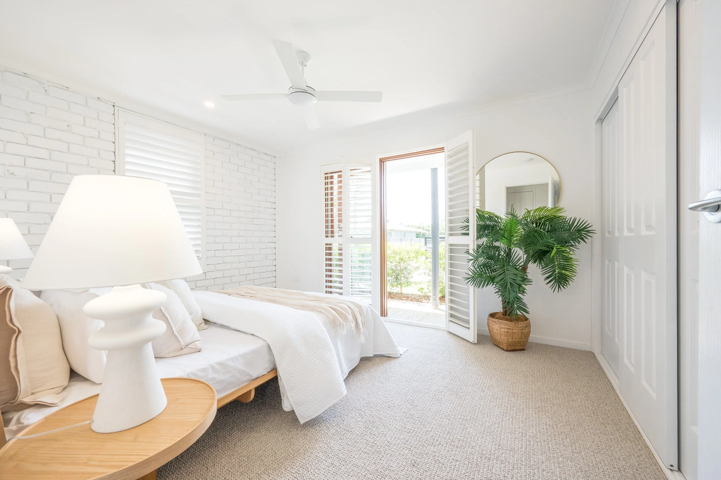 Bright, modern bedroom with white brick wall, large window with white shutters, beige bed with white linens, a wooden side table with a white lamp, and a large potted green plant near a round mirror.