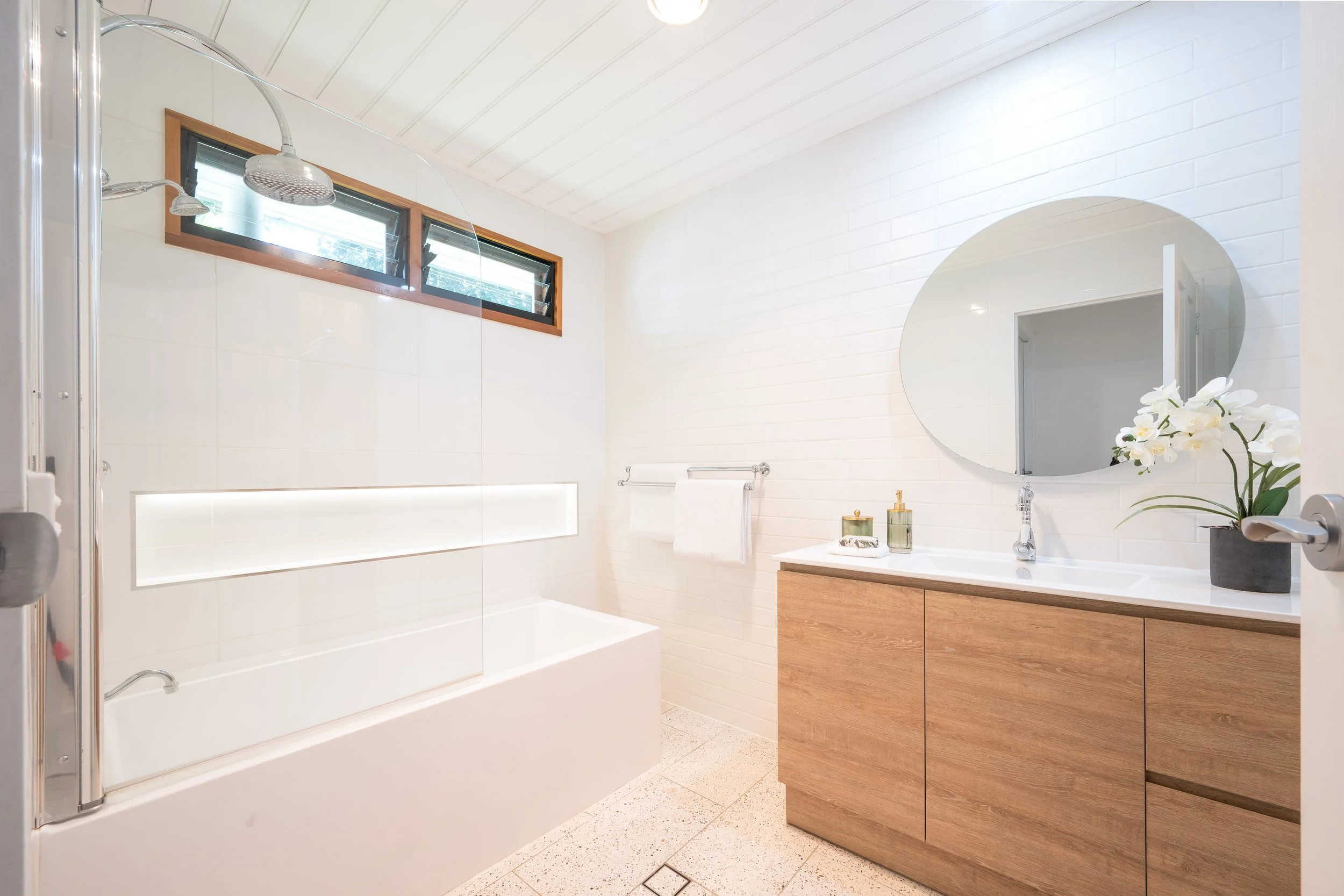 A modern bathroom with a walk-in shower that has glass doors and two heads, a window with three horizontal panes, a white backsplash with a rectangular window, a white oval mirror, a wooden vanity with drawers, a white sink, a brass faucet, and decor