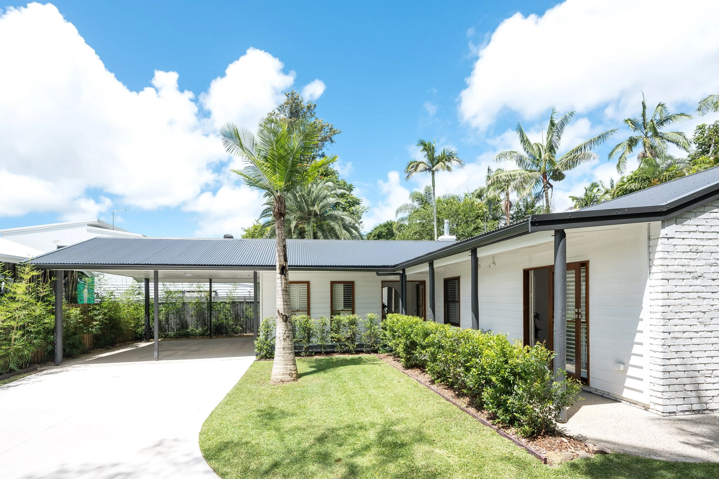 Modern house with white brick walls, gray metal roof, surrounded by green lawn, bushes, palm trees, and a clear blue sky with a few clouds.