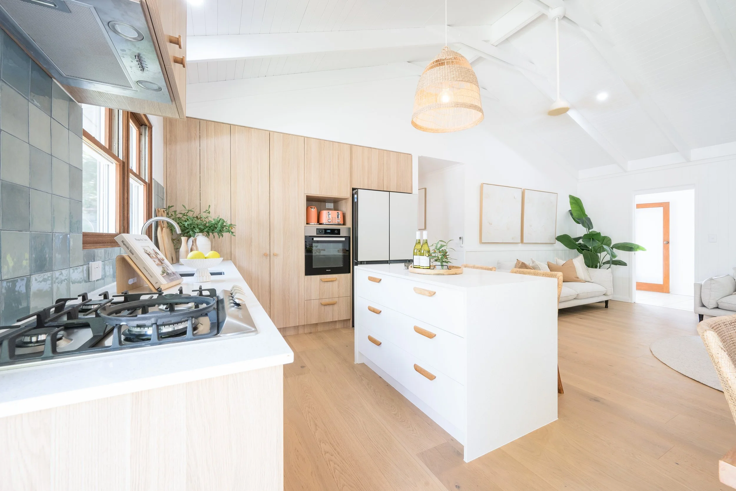 Bright open-concept kitchen and living room with white walls, light wood flooring, and modern decor. The kitchen features a white island with wood details, a gas stove, and a window with natural light. The living area has a white sofa, a large green 