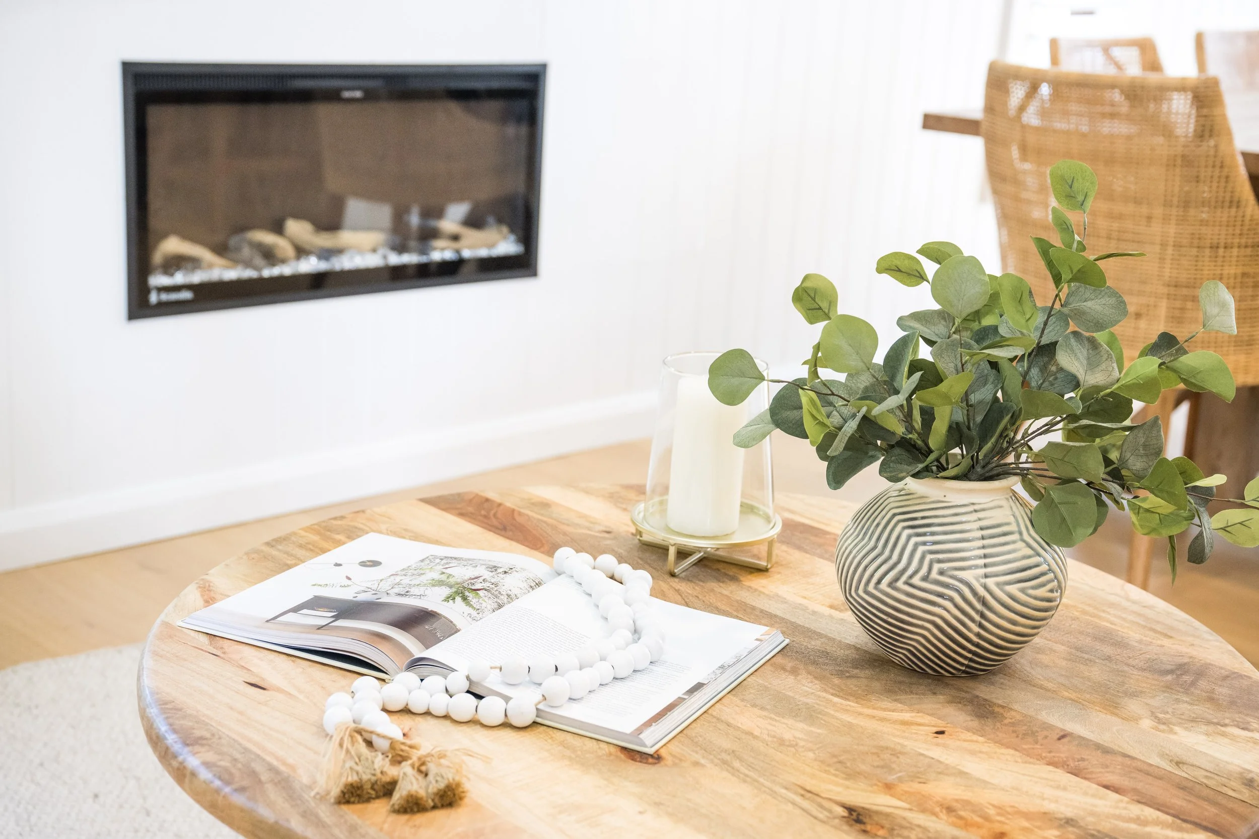 A round wooden table with a striped ceramic vase holding green leafy branches, an open magazine, a white candle in a glass holder, and a white beaded decorative item. In the background, a flat-screen TV, a rattan chair, and a green plant are visible 