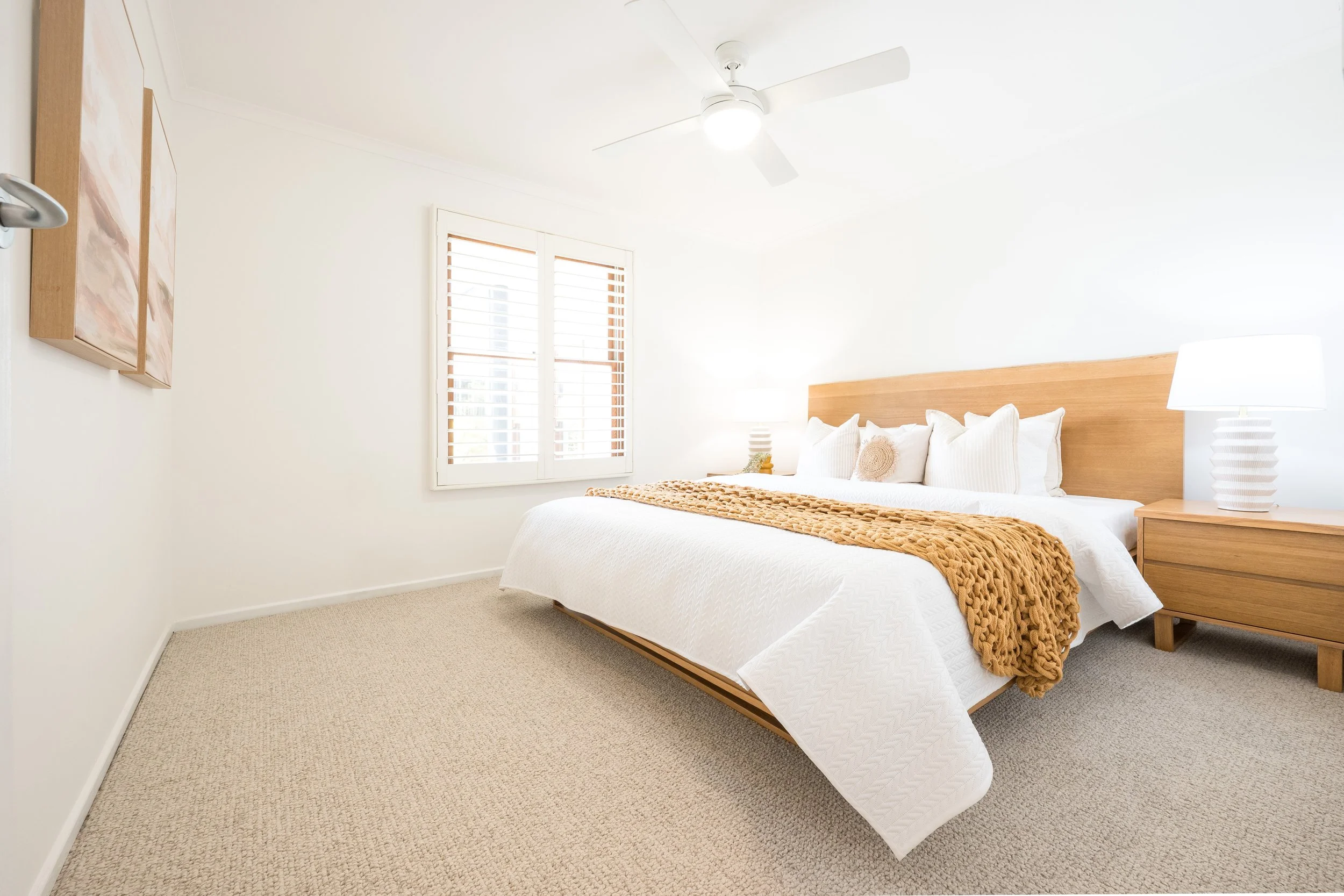 Bright bedroom with a large bed, white bedding, a brown knit blanket, wooden nightstand with a white lamp, a window with white shutters, a ceiling fan, and a wall hanging.