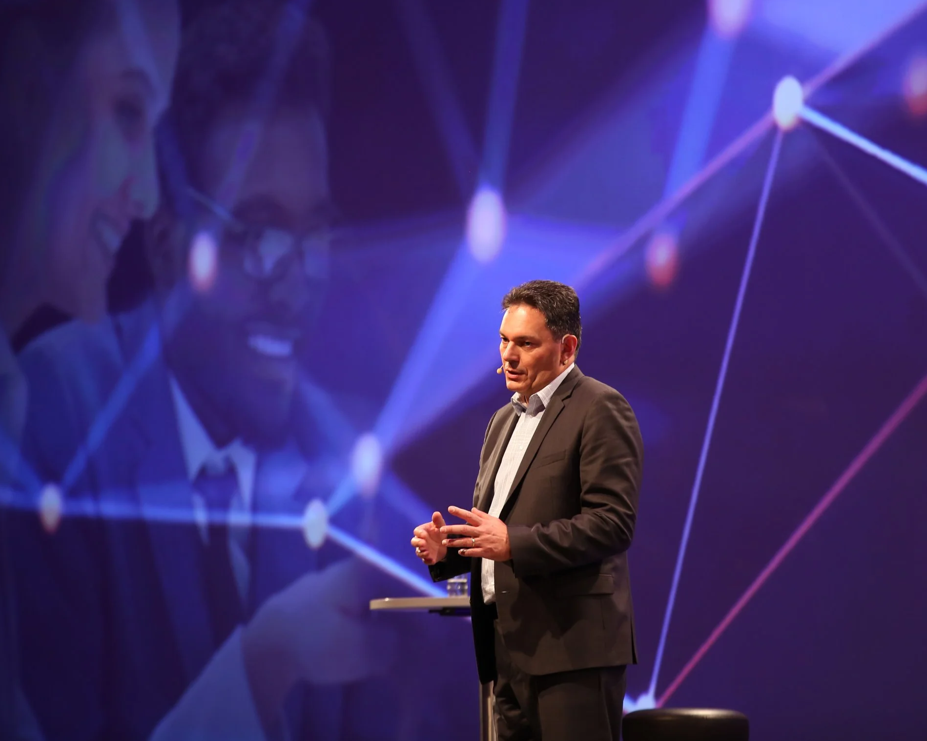 A man in a suit giving a presentation on stage with a dark purple background and connecting line graphics.