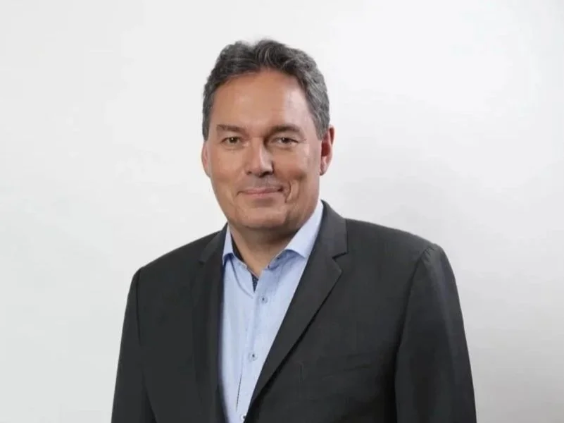 Man in a dark suit and light blue shirt standing against a plain white background.